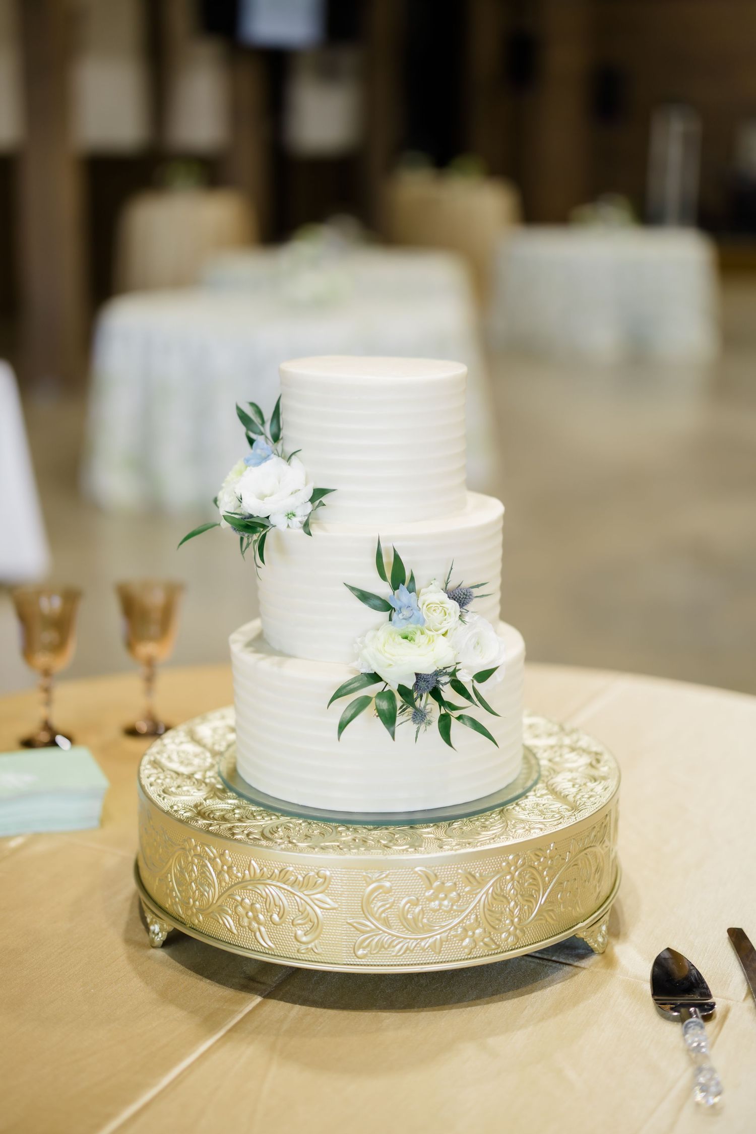 Three-tier white wedding cake with floral accents on a gold stand at a reception.