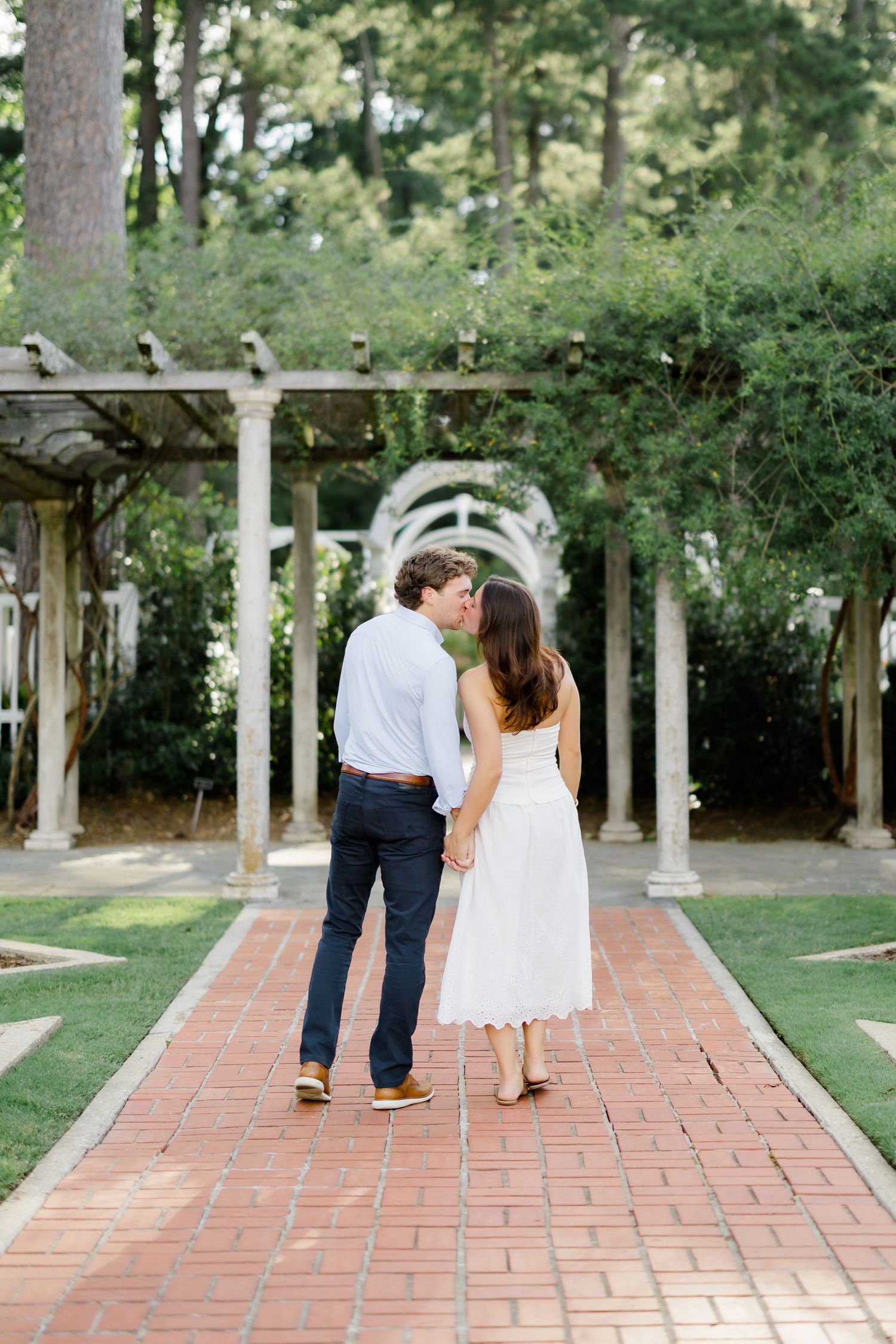 A couple shares a kiss while holding hands on a brick garden path lined with white columns and lush greenery.