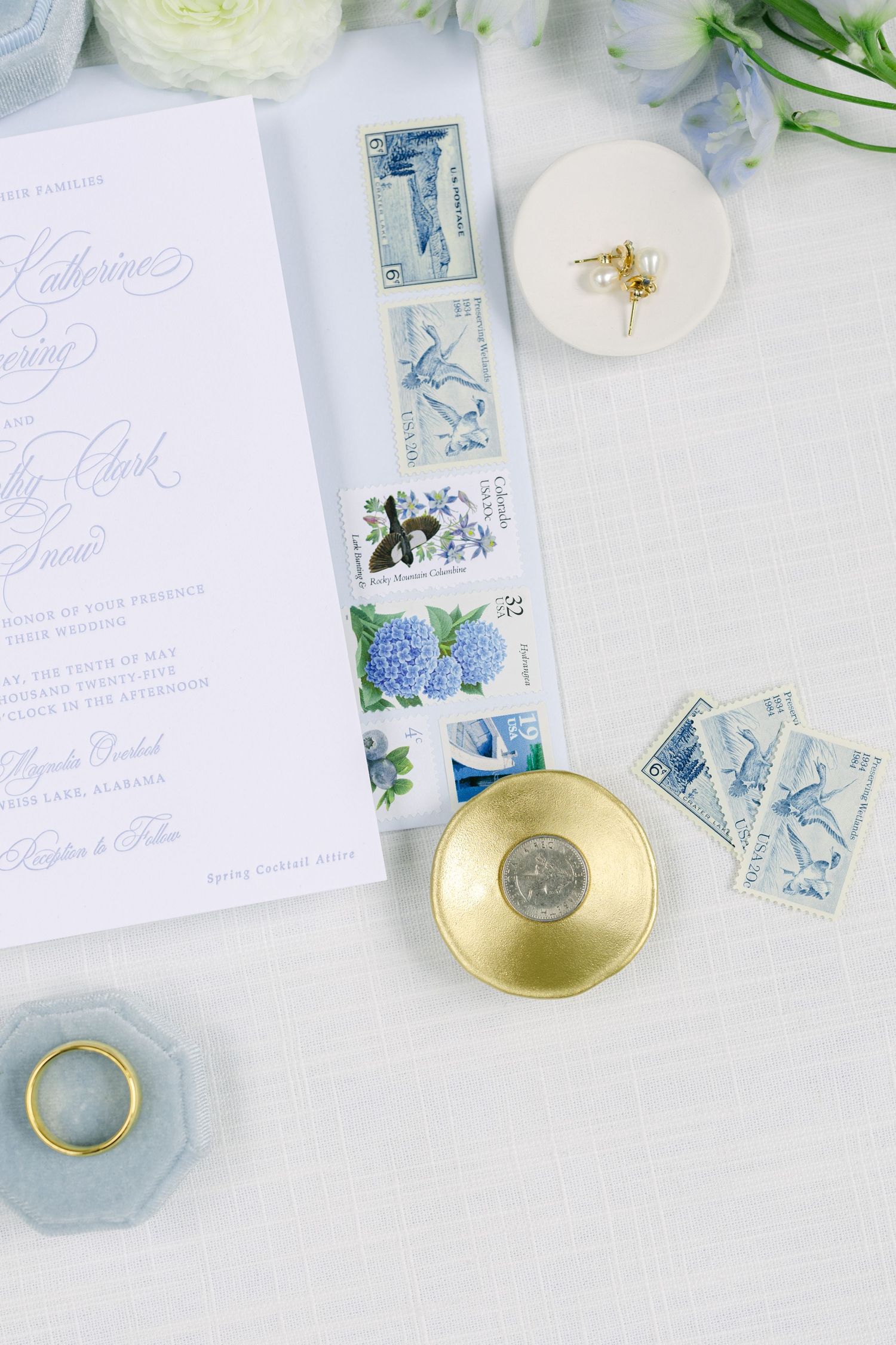 Elegant wedding invitations with blue vintage floral stamps and gold ring dishes arranged on white background. Sixpence wedding tradition