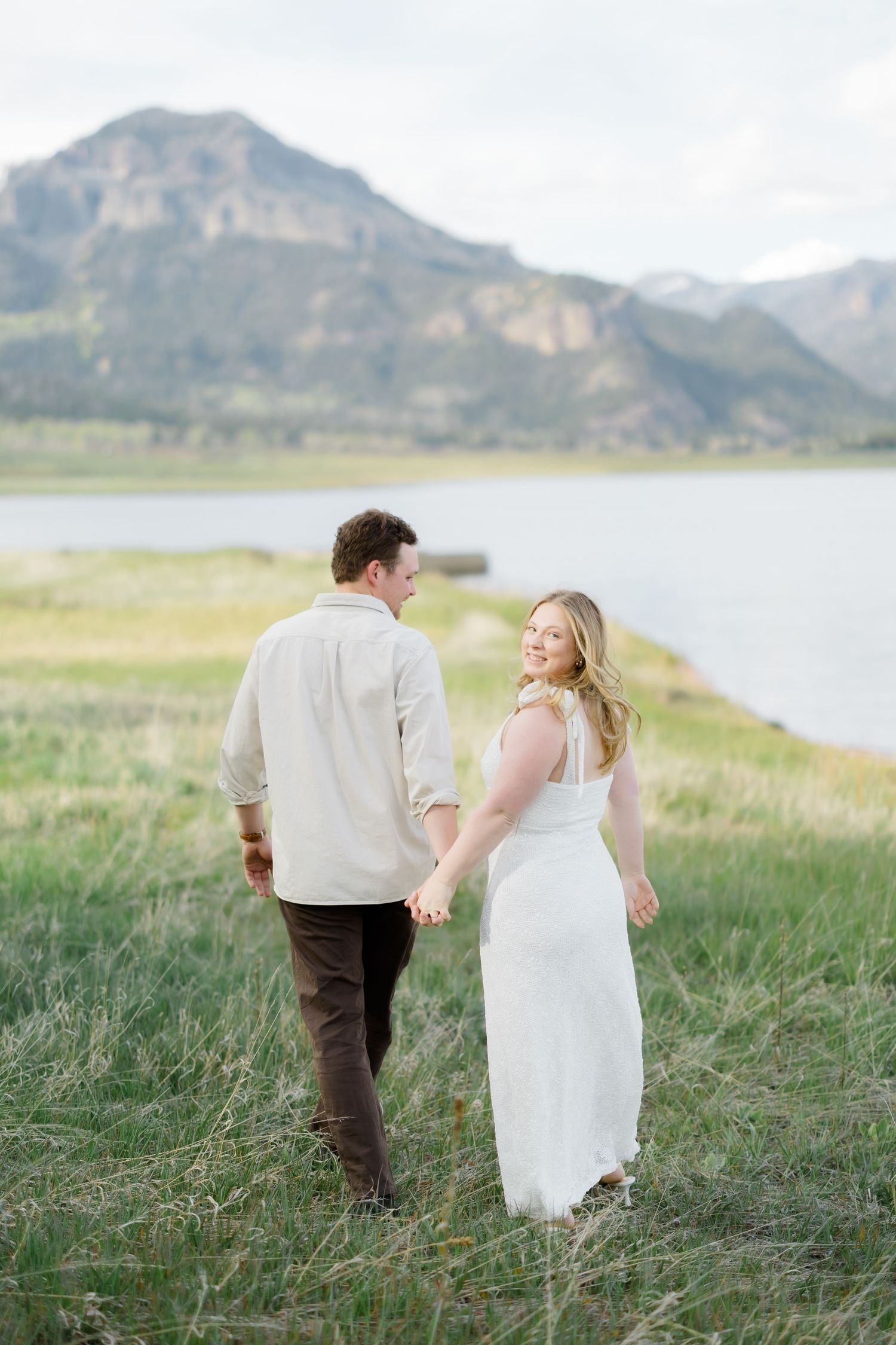 Engaged couple walking hand in hand through green meadow with mountain lake backdrop.