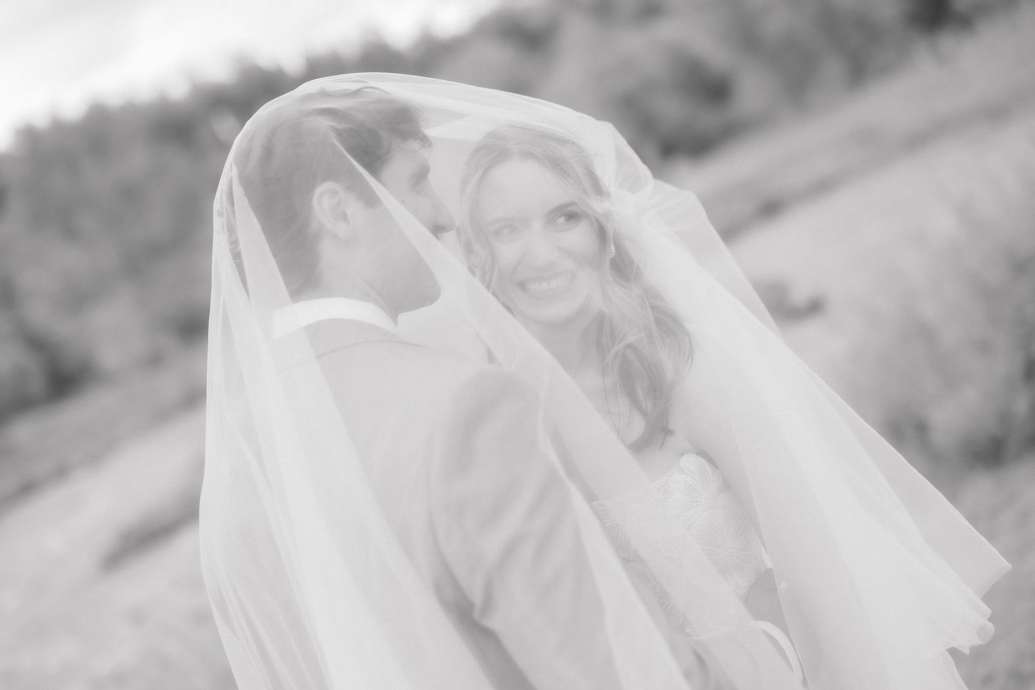 Black and white wedding photo of bride and groom sharing a romantic moment under a flowing veil outdoors.