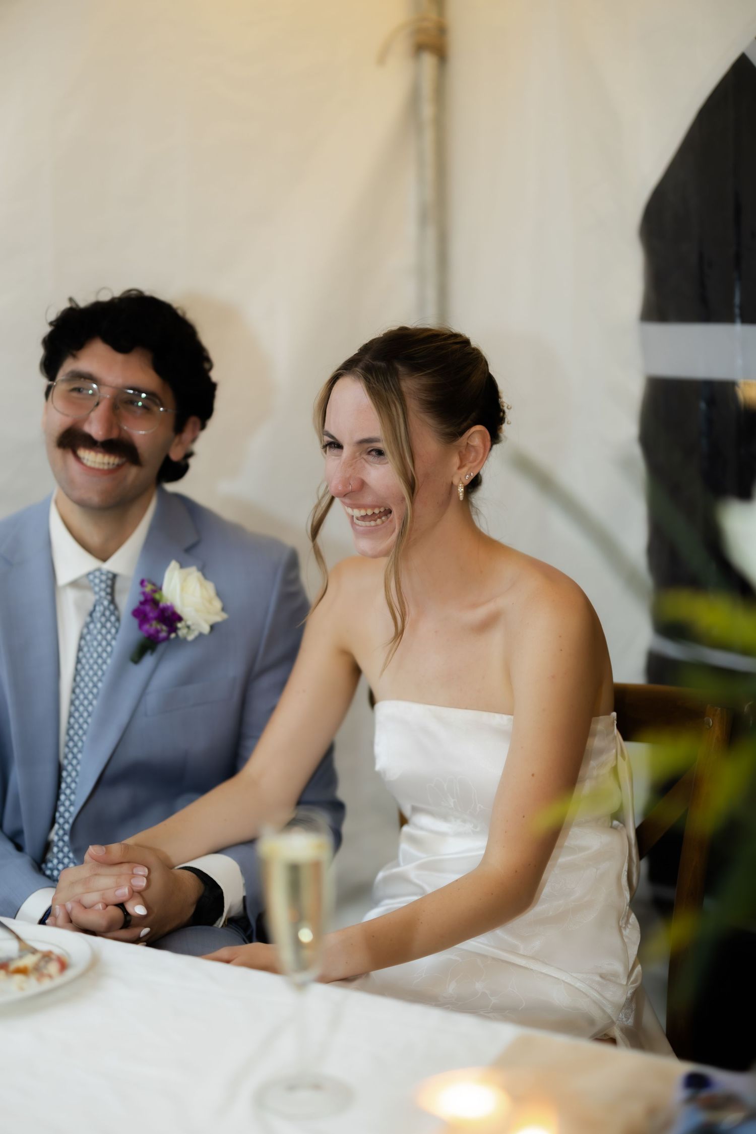 Smiling bride in strapless gown and groom in gray suit holding hands at wedding reception table.