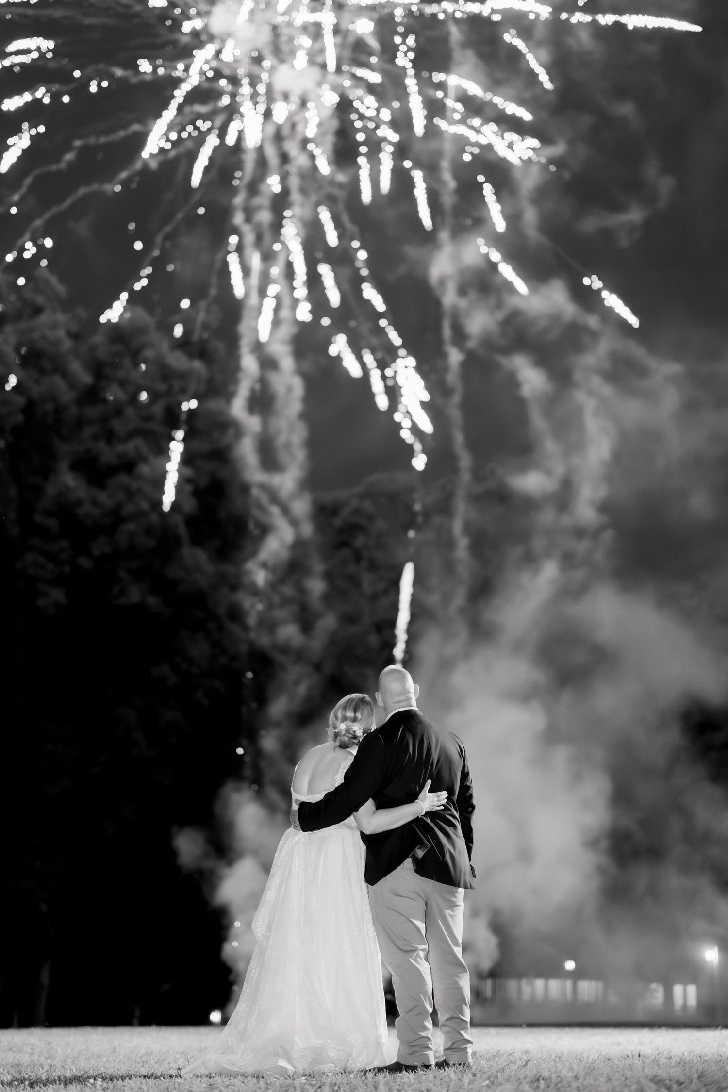 Black and white photo of a bride and groom embracing while watching fireworks light up the night sky.