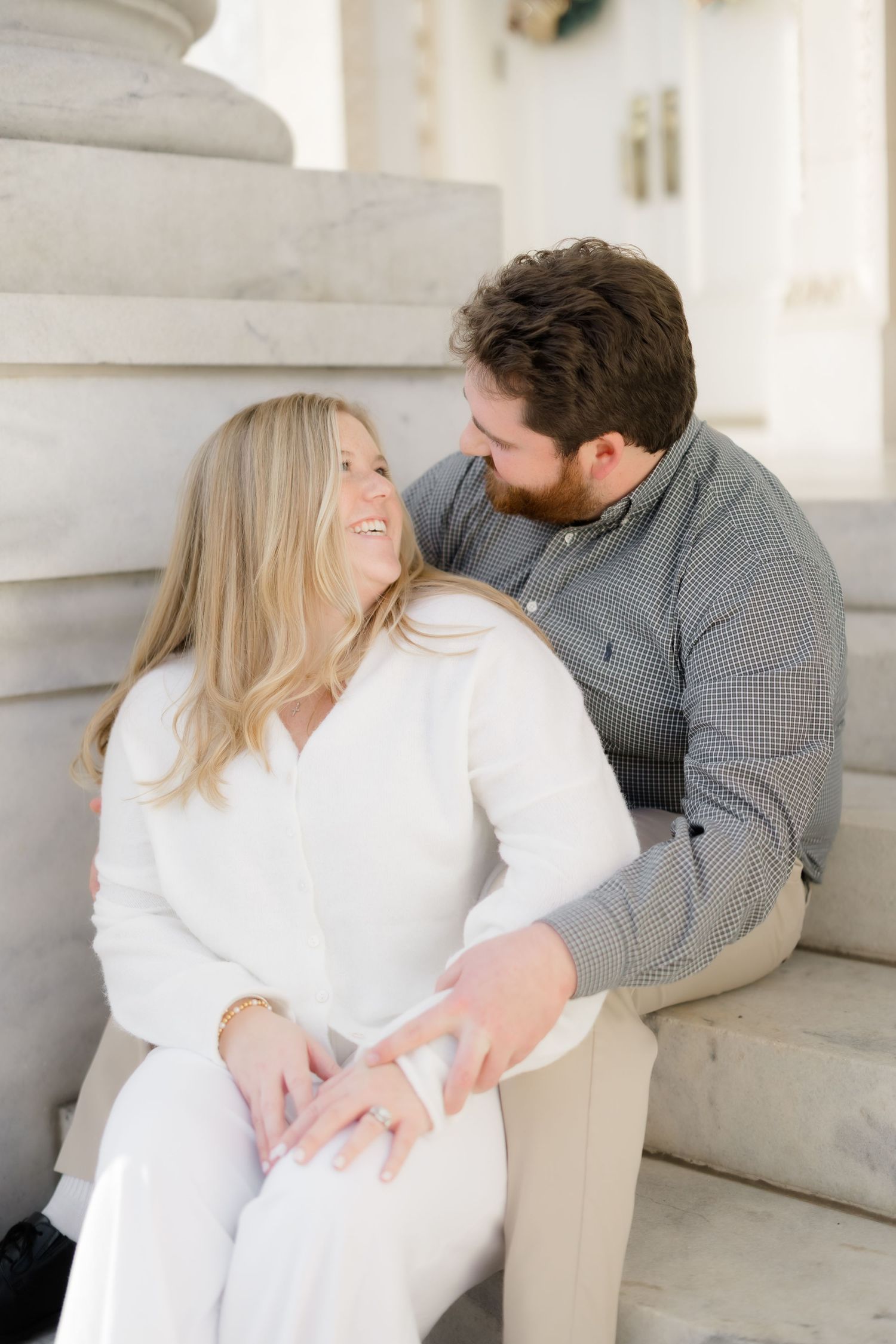 Happy couple sits on marble steps, woman in white laughing as man leans in close, engagement photo session.