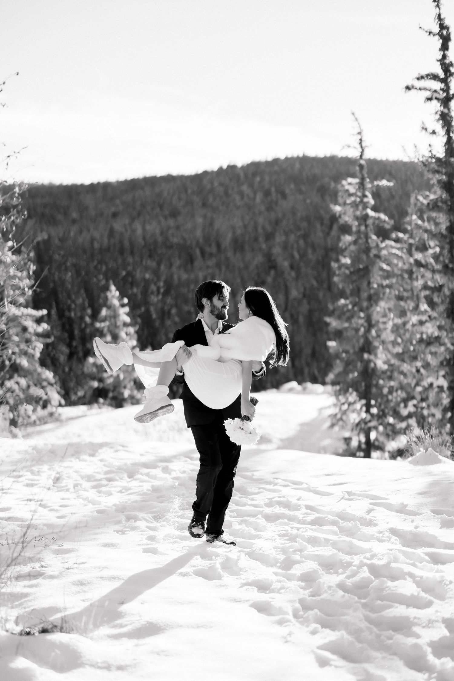 Black and white photo of groom lifting bride in snowy mountain forest during winter elopement.