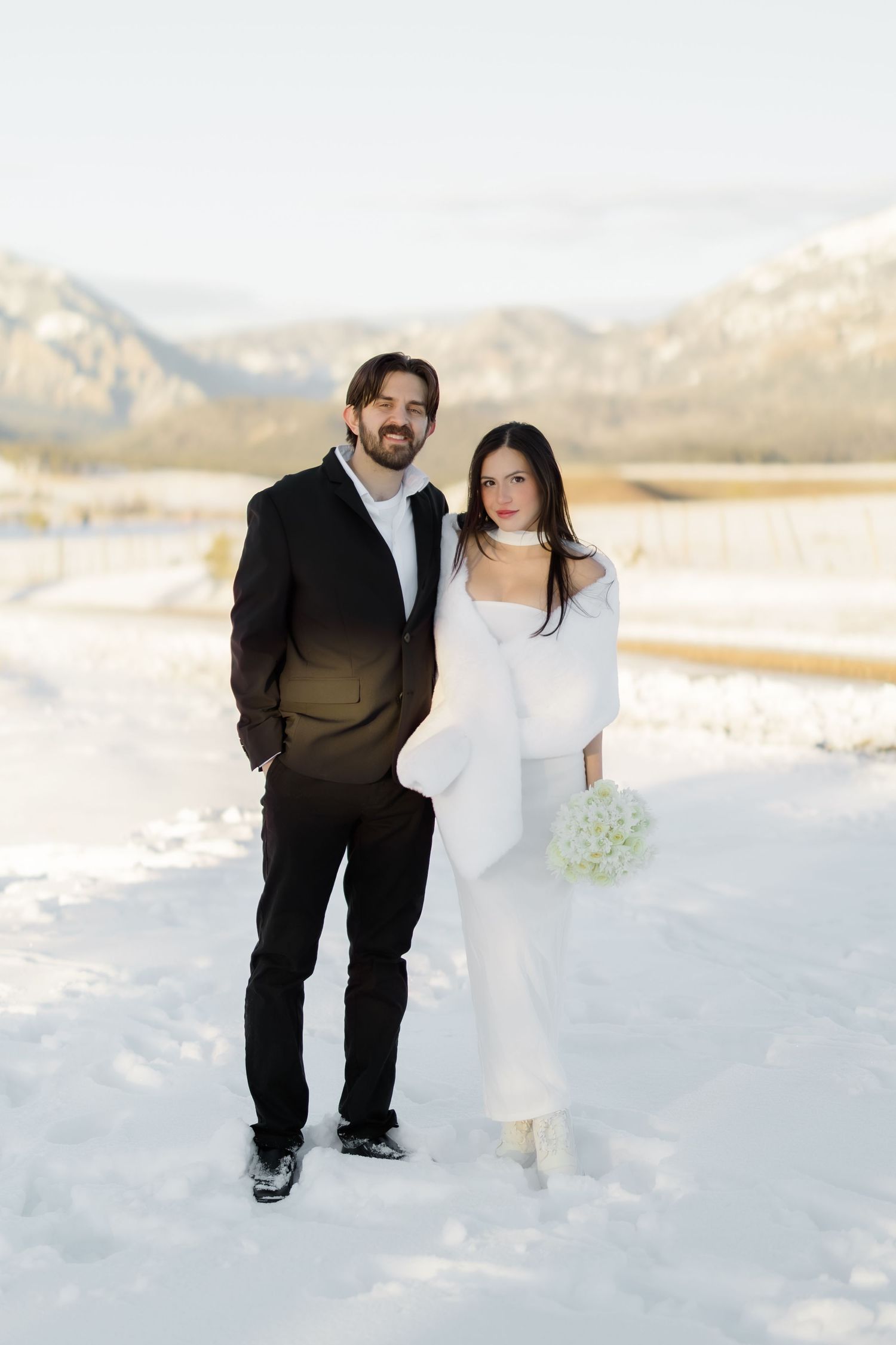 Smiling bride and groom posing together in snow with mountain backdrop during winter wedding shoot.