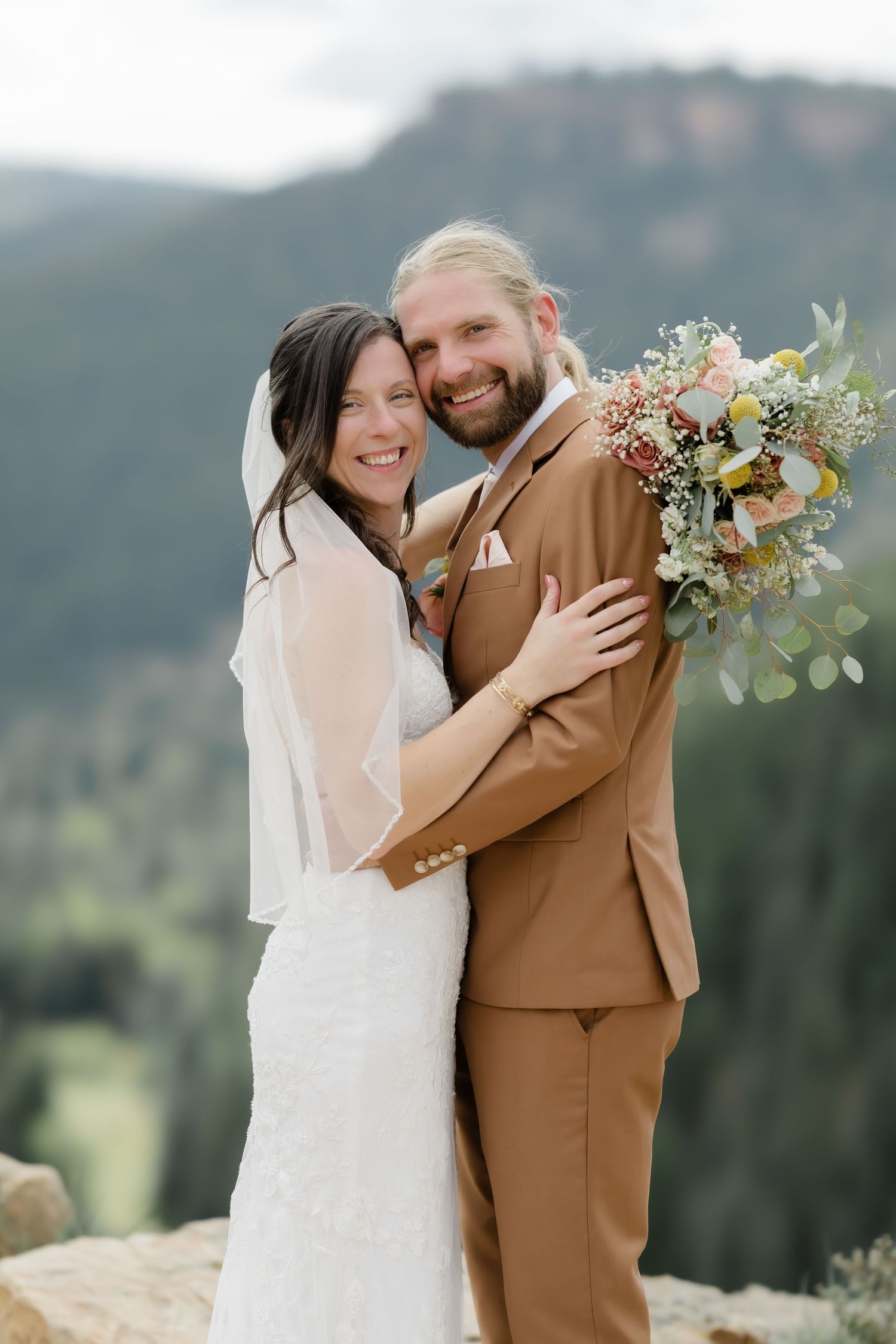 Smiling bride and groom embrace outdoors with mountain views, she wears a veil, he holds a colorful bouquet in a tan suit.