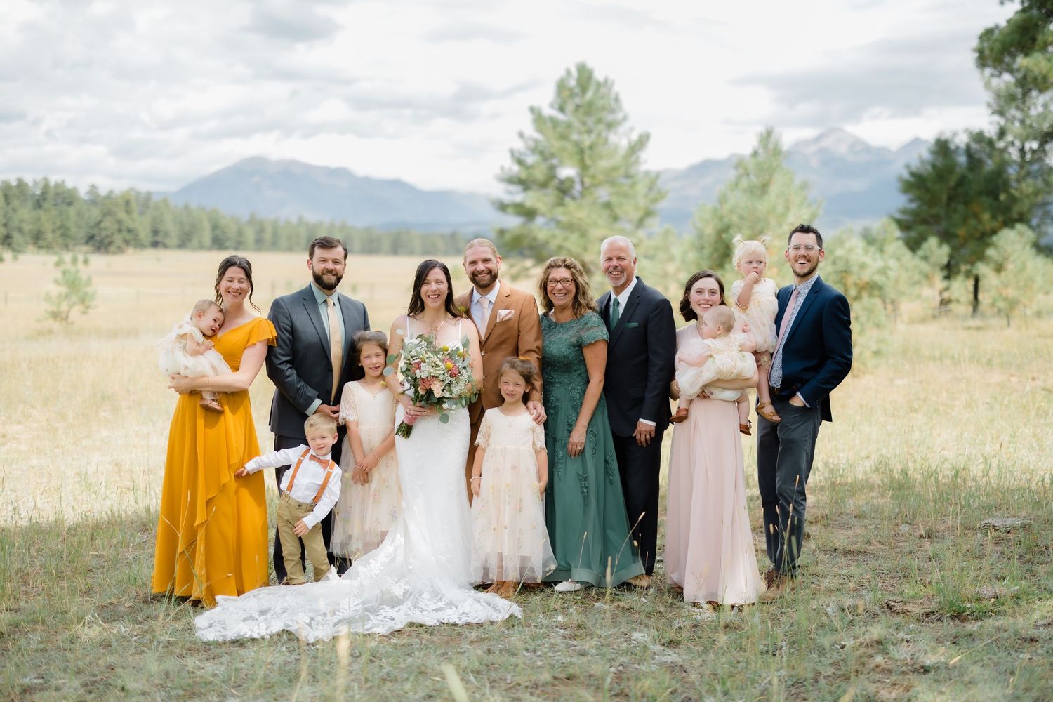 Wedding party poses outdoors in mountain meadow, bride in white gown with family in yellow, green, and blush attire.
