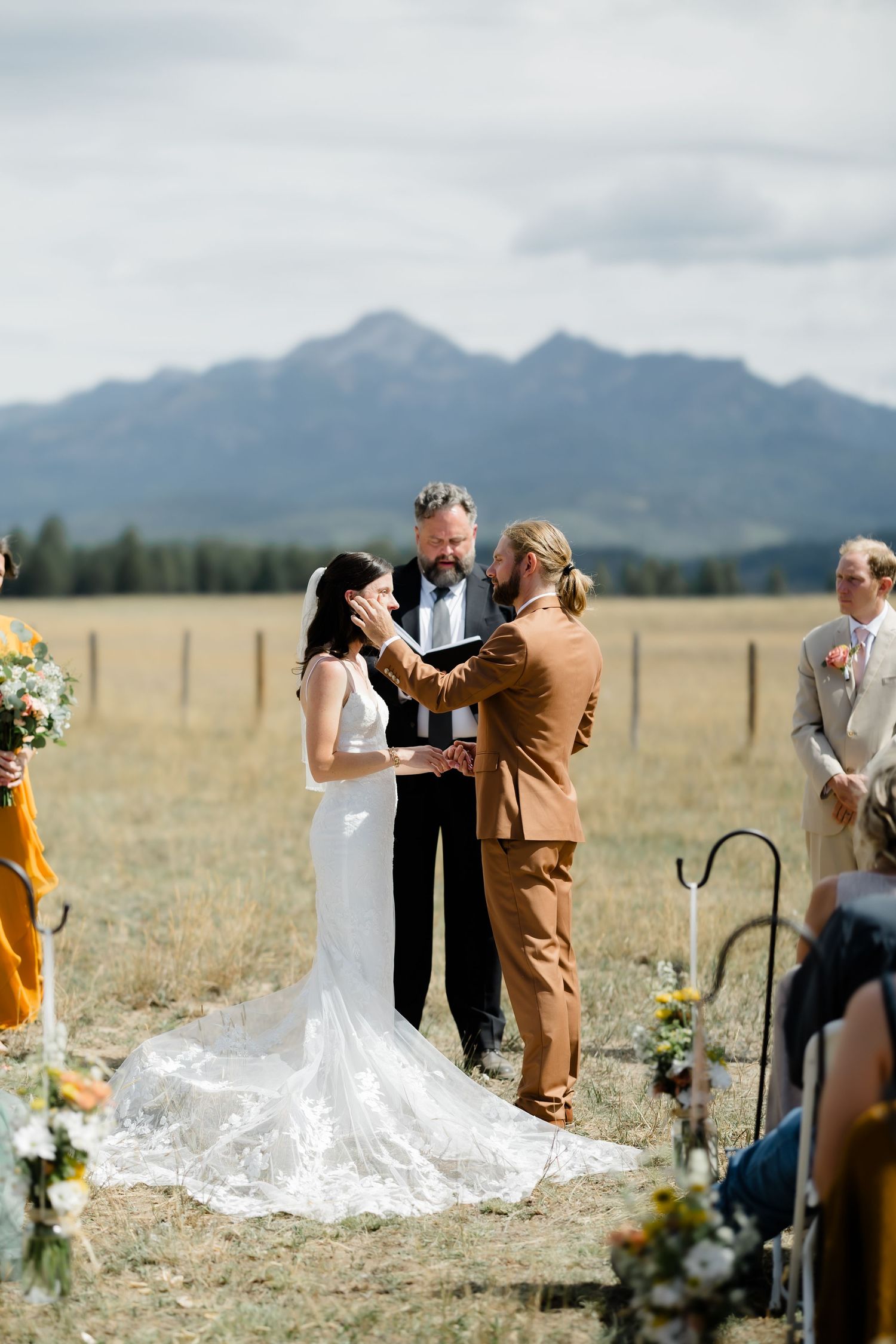 Outdoor wedding ceremony in a mountain meadow with bride in white gown and groom in tan suit exchanging vows.