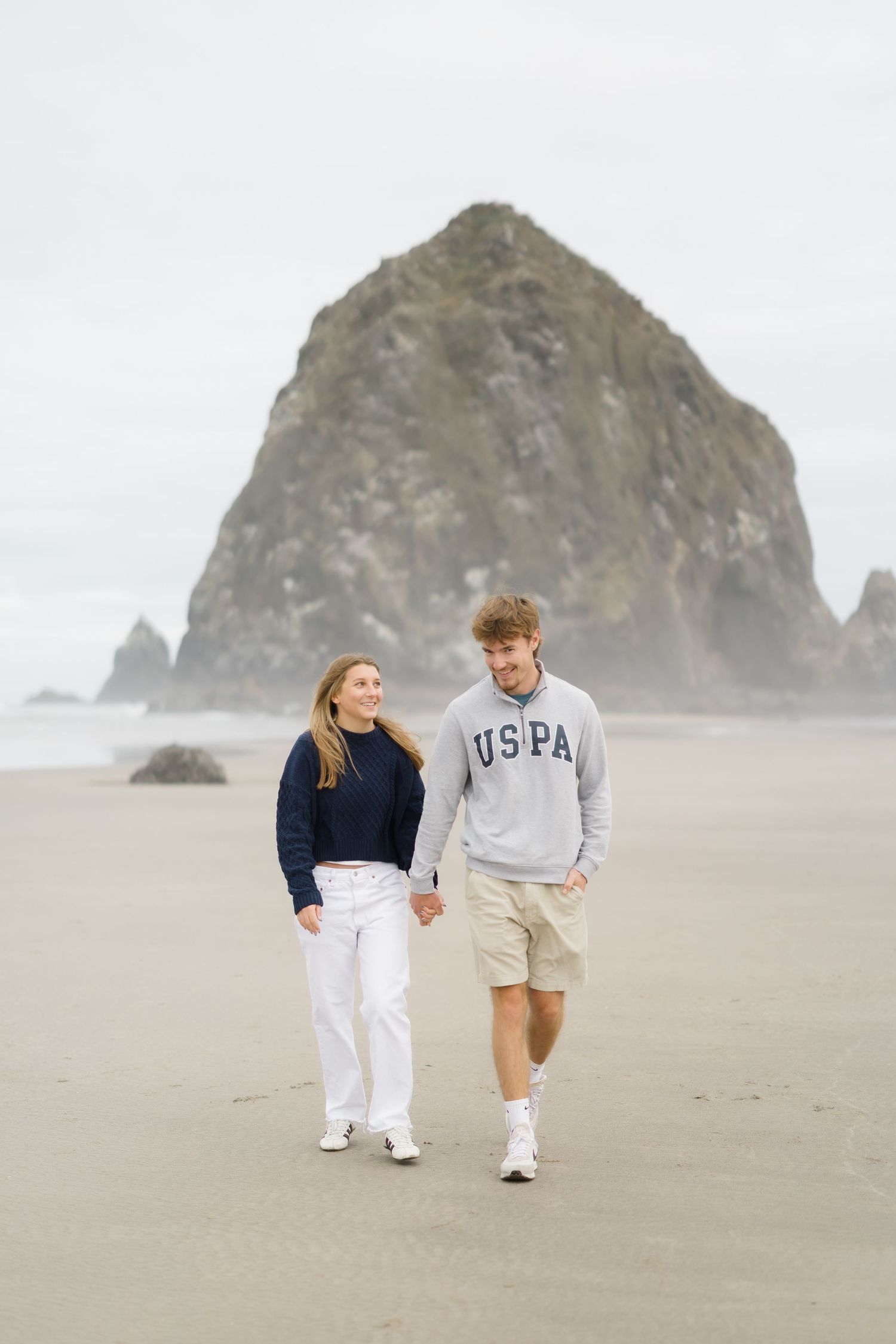 Couple walking hand in hand on a sandy beach with Haystack Rock in the background on a cloudy day.