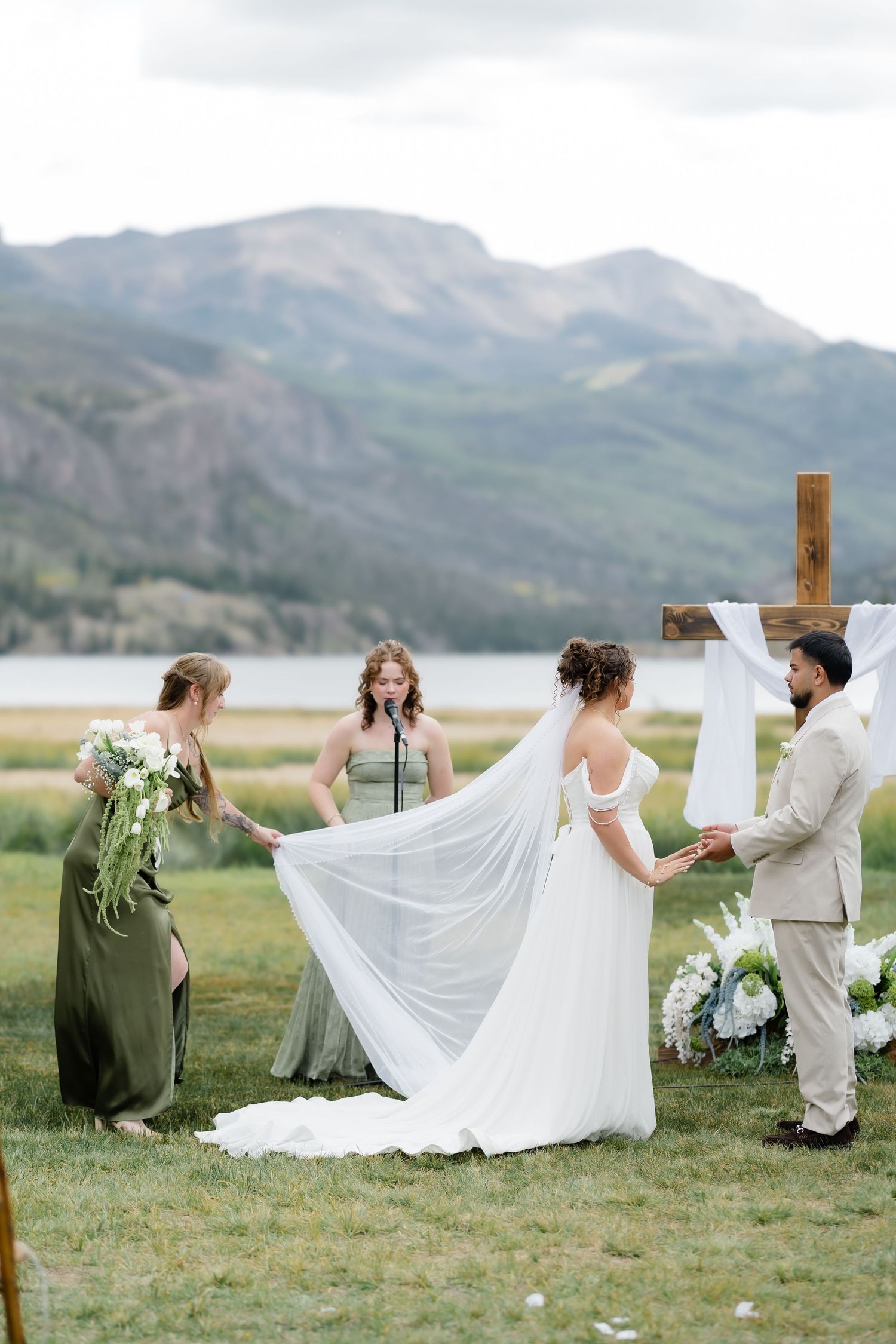 Bride in white gown with veil at outdoor mountain lake wedding ceremony with wooden cross altar.