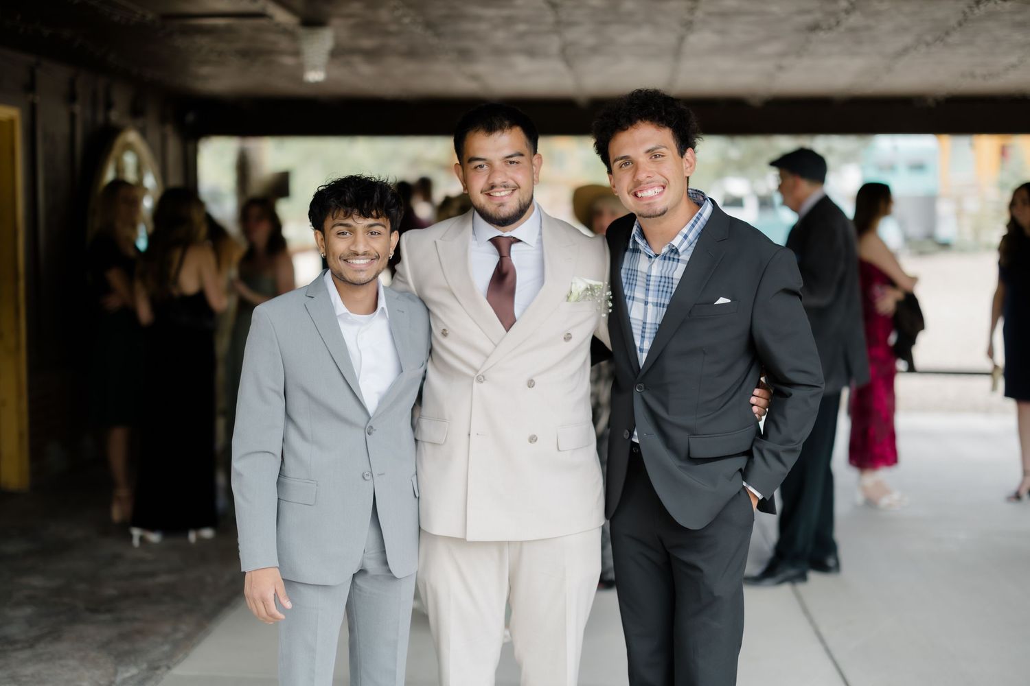 Three young men in suits smiling together at a formal event under a covered outdoor venue.