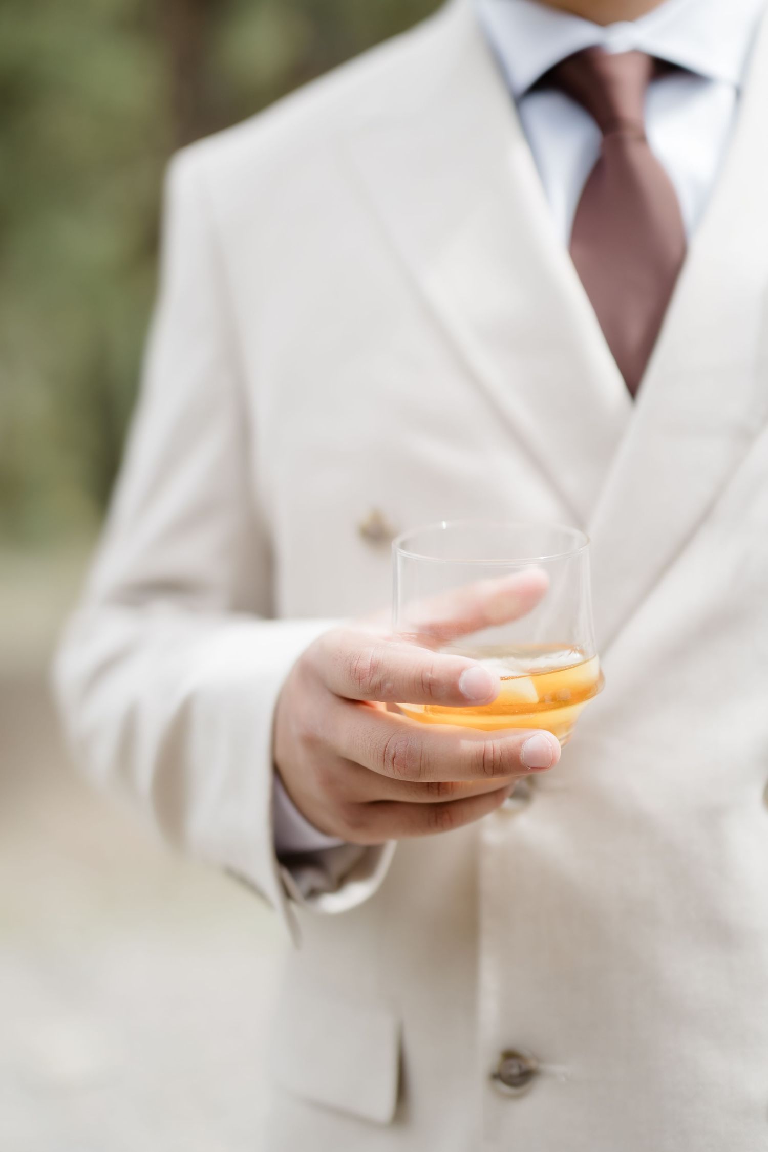 Man in beige suit holding a glass of whiskey with ice, wearing a burgundy tie.