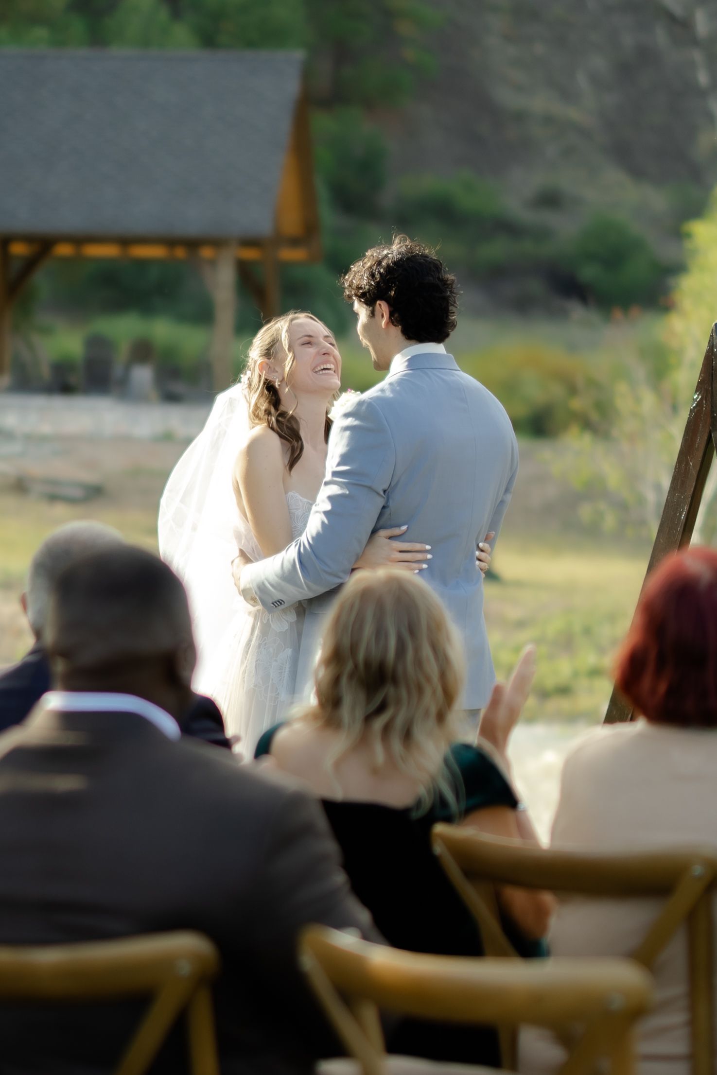 Bride and groom share a joyful moment during outdoor wedding ceremony with guests seated in wooden chairs.