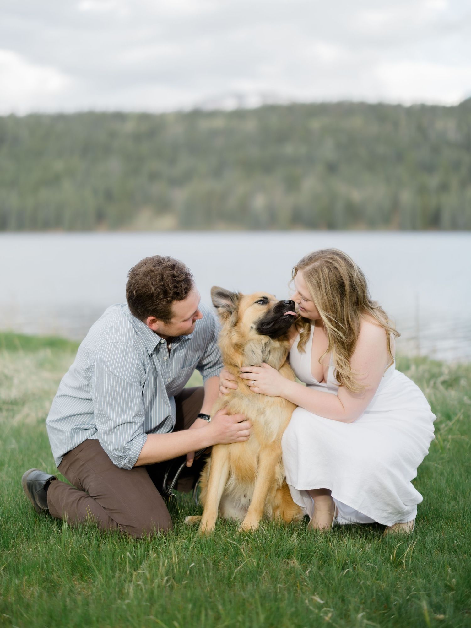 Couple kneeling on grass with German Shepherd dog near lakeside, sharing a joyful moment together.