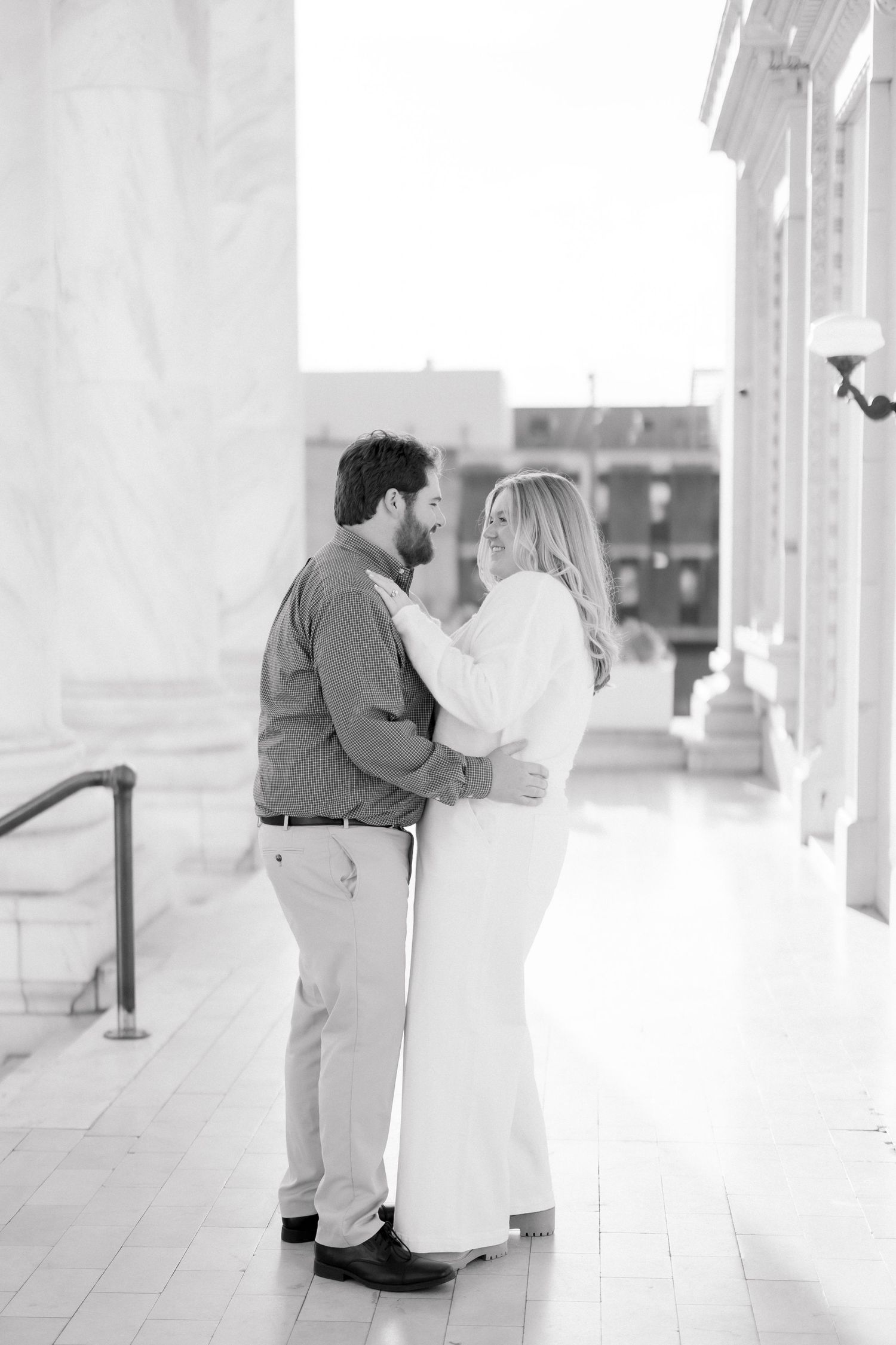 Black and white photo of an engaged couple embracing and gazing at each other in a marble hallway.