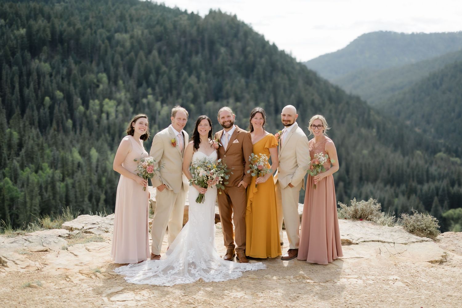Wedding party poses on rocky mountain overlook, bride in white, groom in rust suit, attendants in blush and tan.