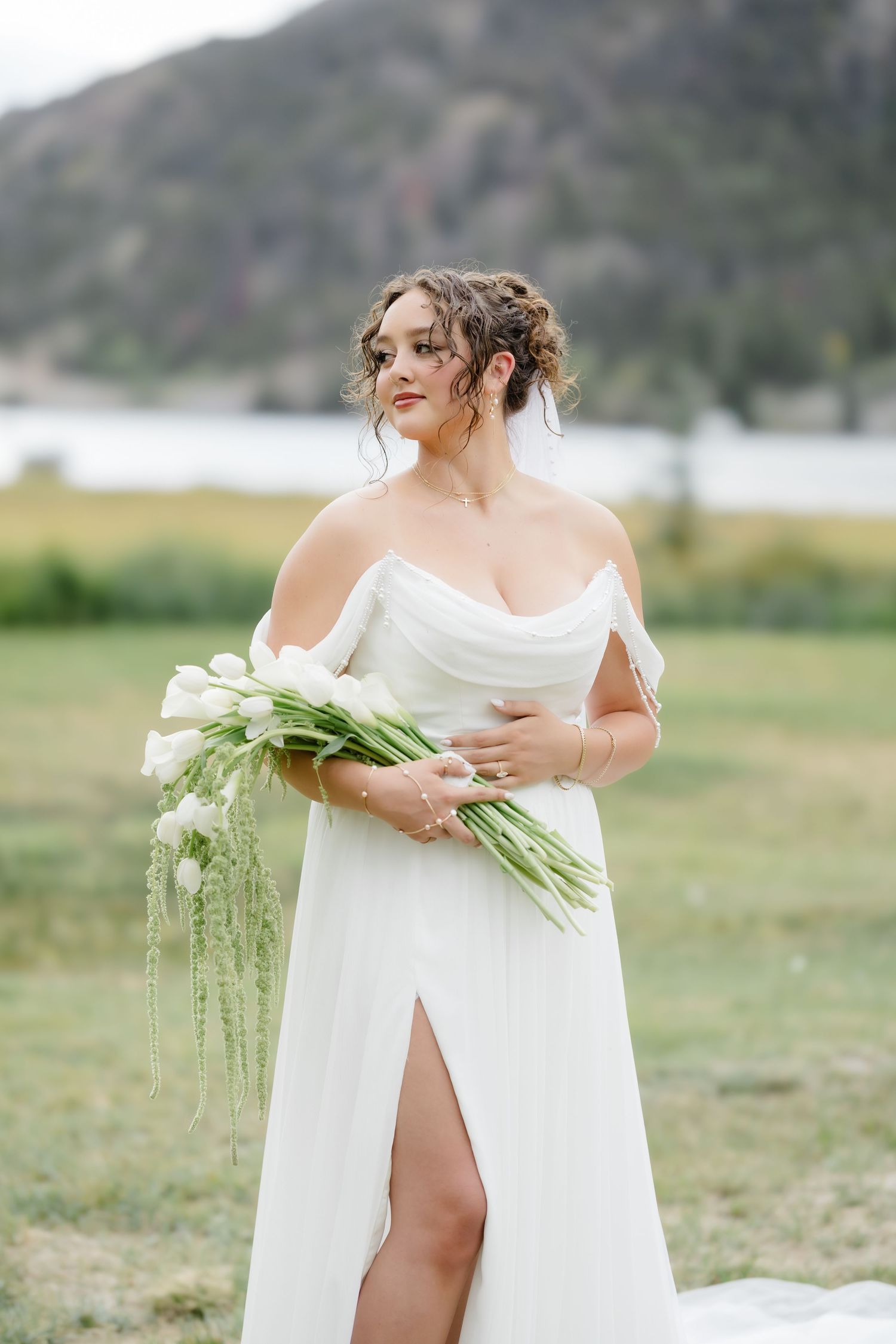 Bride in white off-shoulder gown holds white tulip bouquet outdoors near mountain lake.