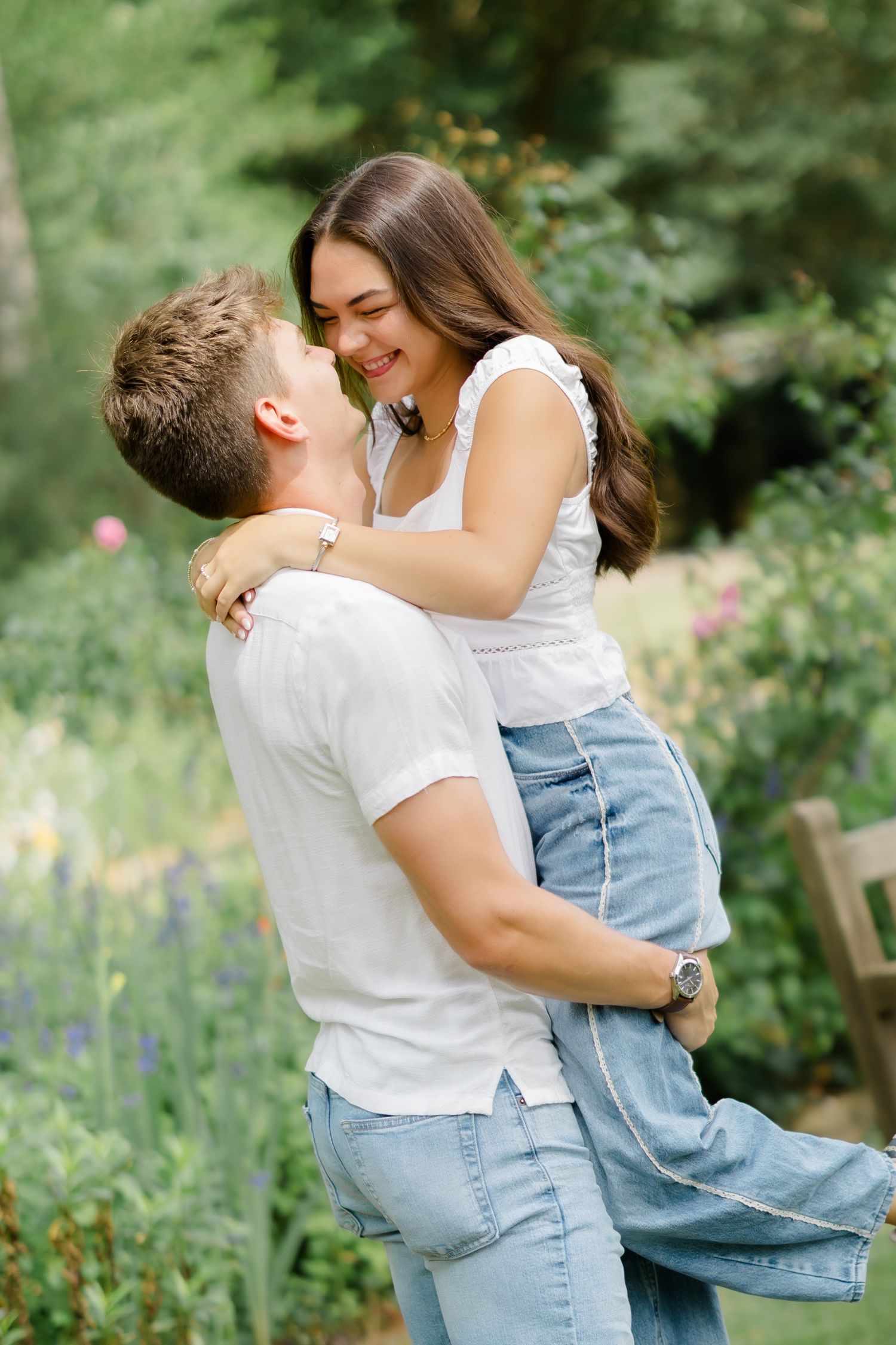 A couple in white tops and jeans embrace outdoors, the man lifting the smiling woman in a lush green garden.