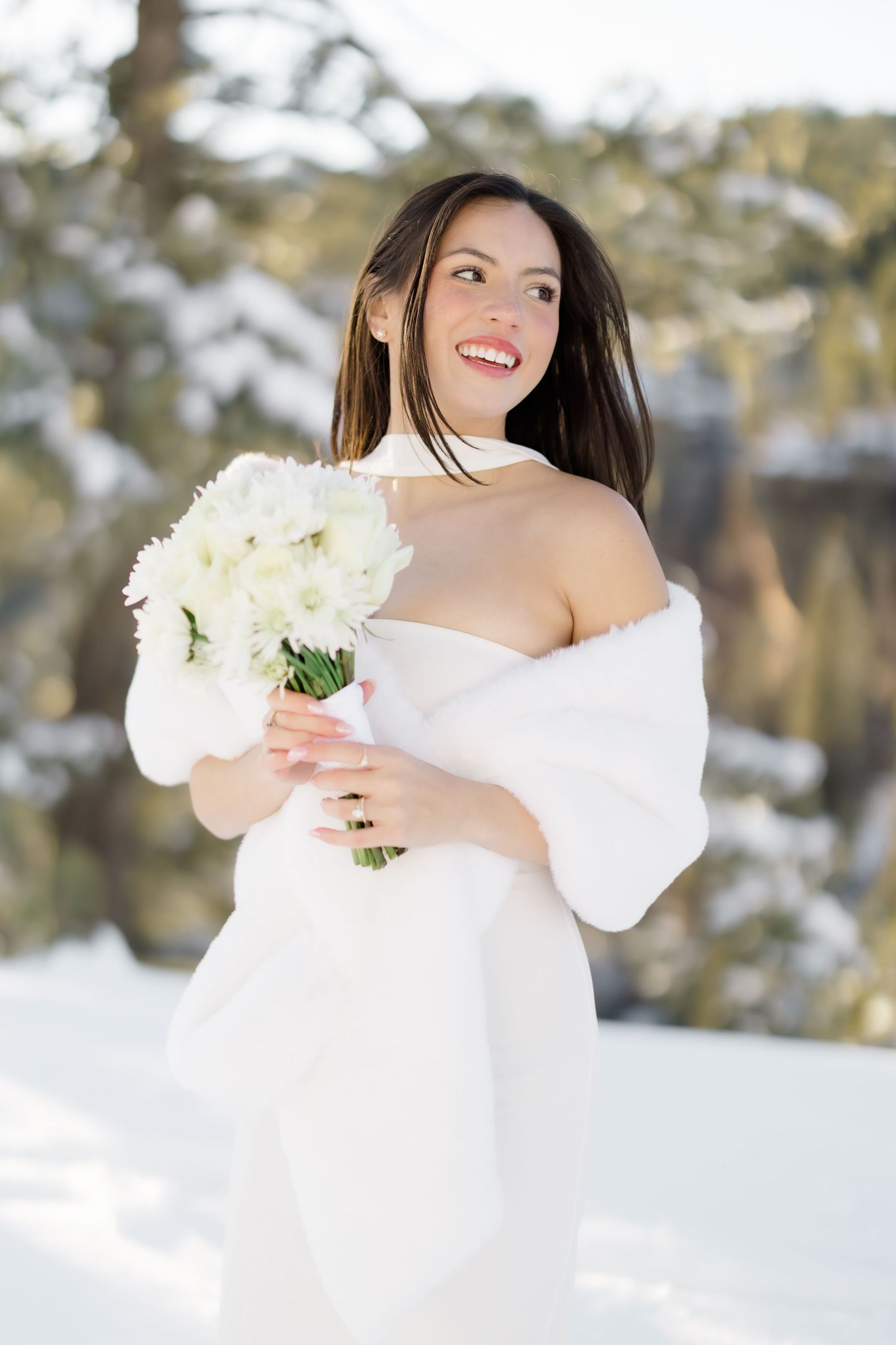 Smiling bride in white gown and fur shawl holds white flower bouquet in winter snow.