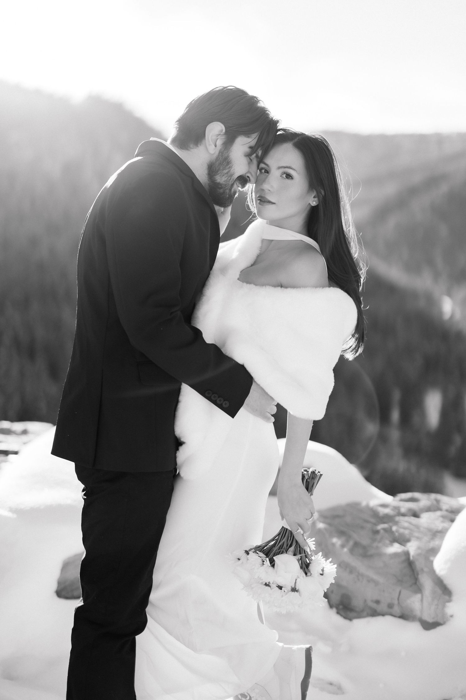 Black and white photo of groom embracing bride in fur wrap with snowy mountain backdrop.