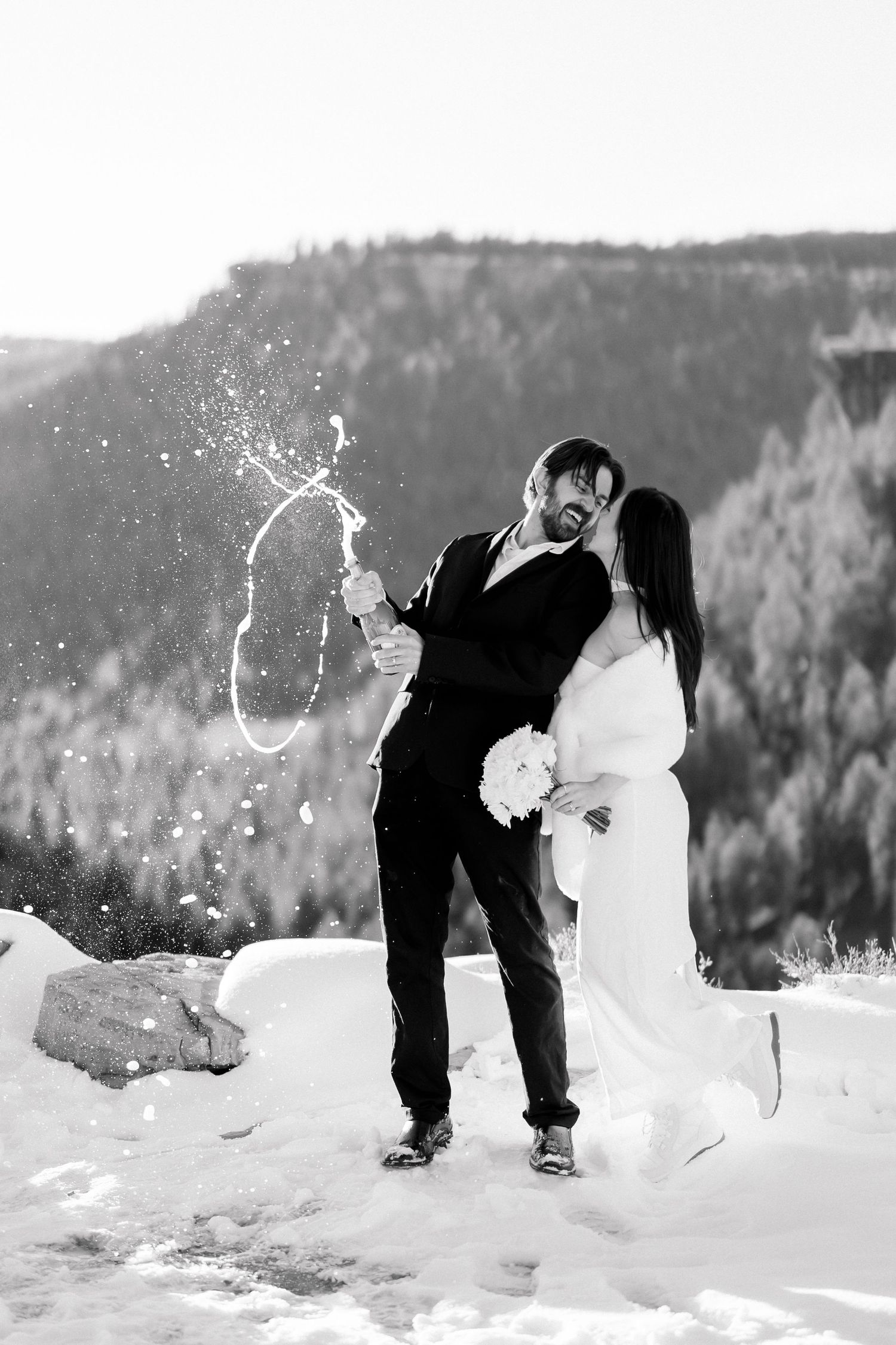 Black and white photo of newlyweds celebrating with champagne spray in a snowy mountain setting.
