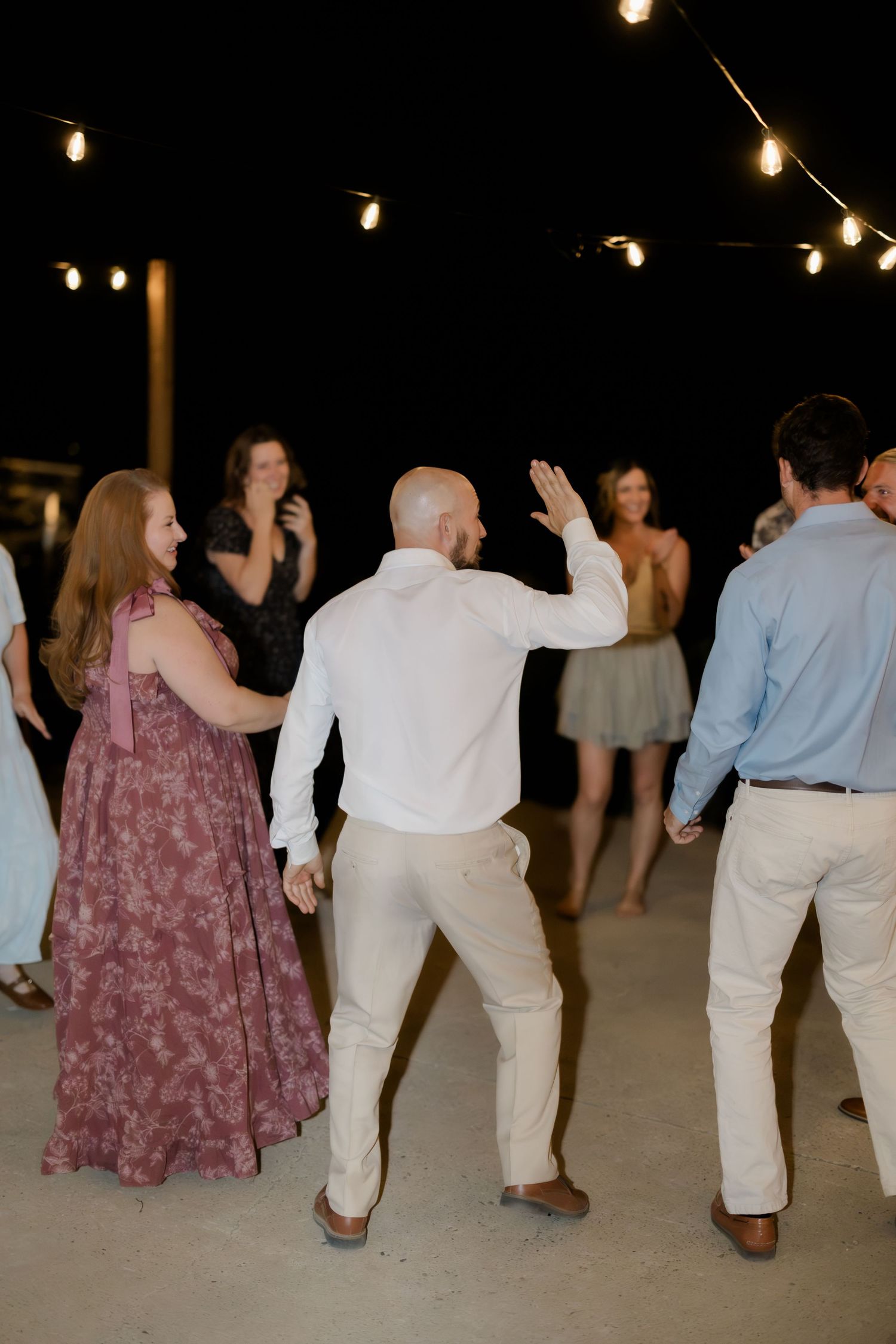 Guests dancing outdoors at a wedding reception under string lights at night, wearing formal attire.