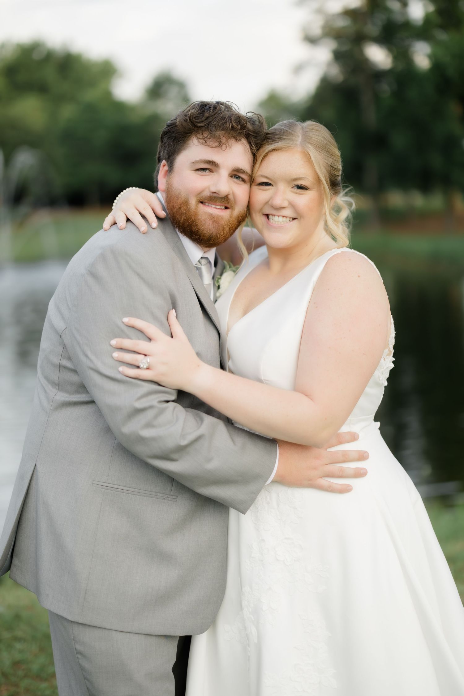 A smiling bride and groom embrace outdoors near a pond, she in a white gown and he in a gray suit.