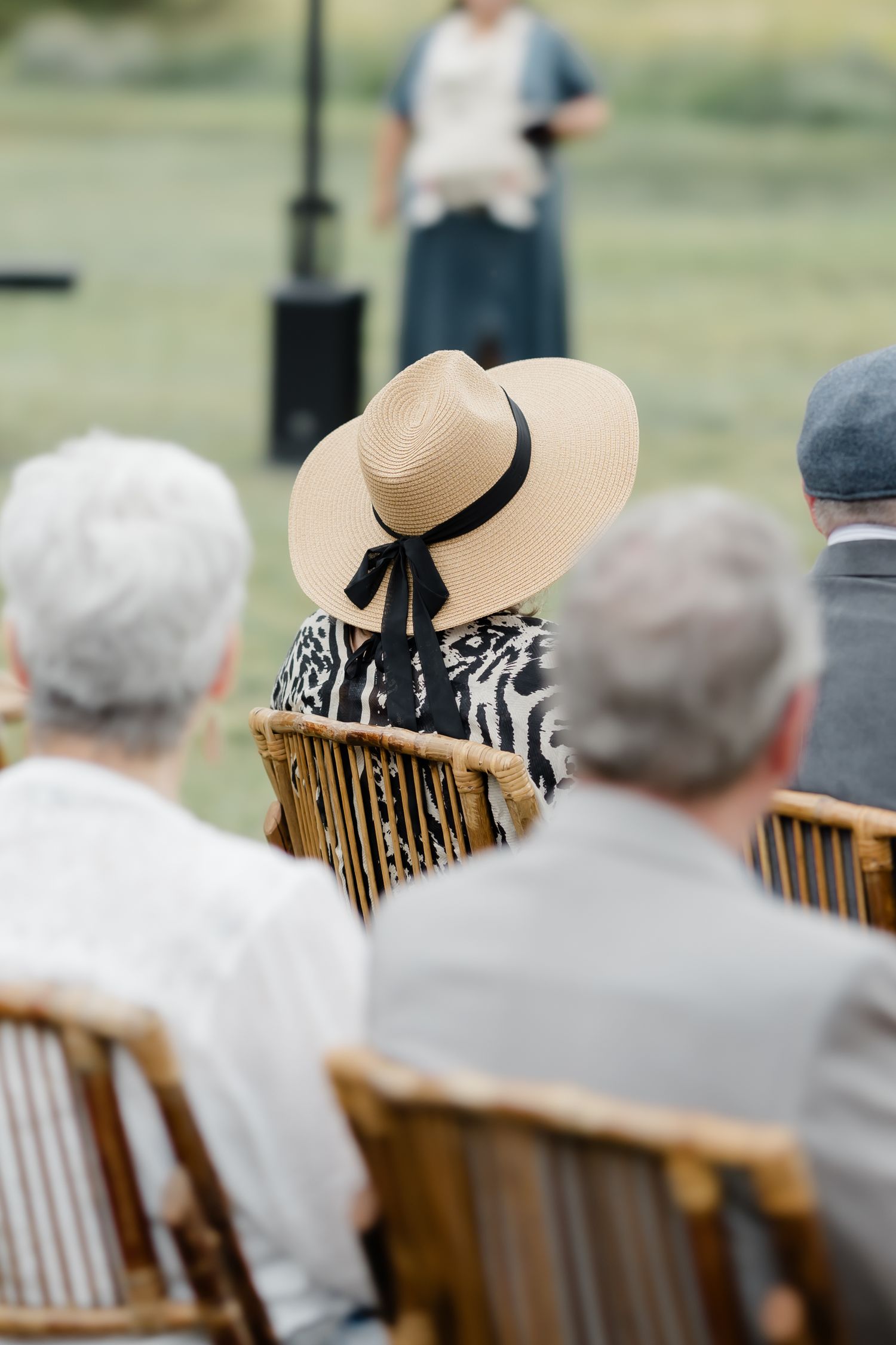 Guests seated outdoors at an elegant event, woman in wide-brim straw hat with black ribbon, speaker in background.
