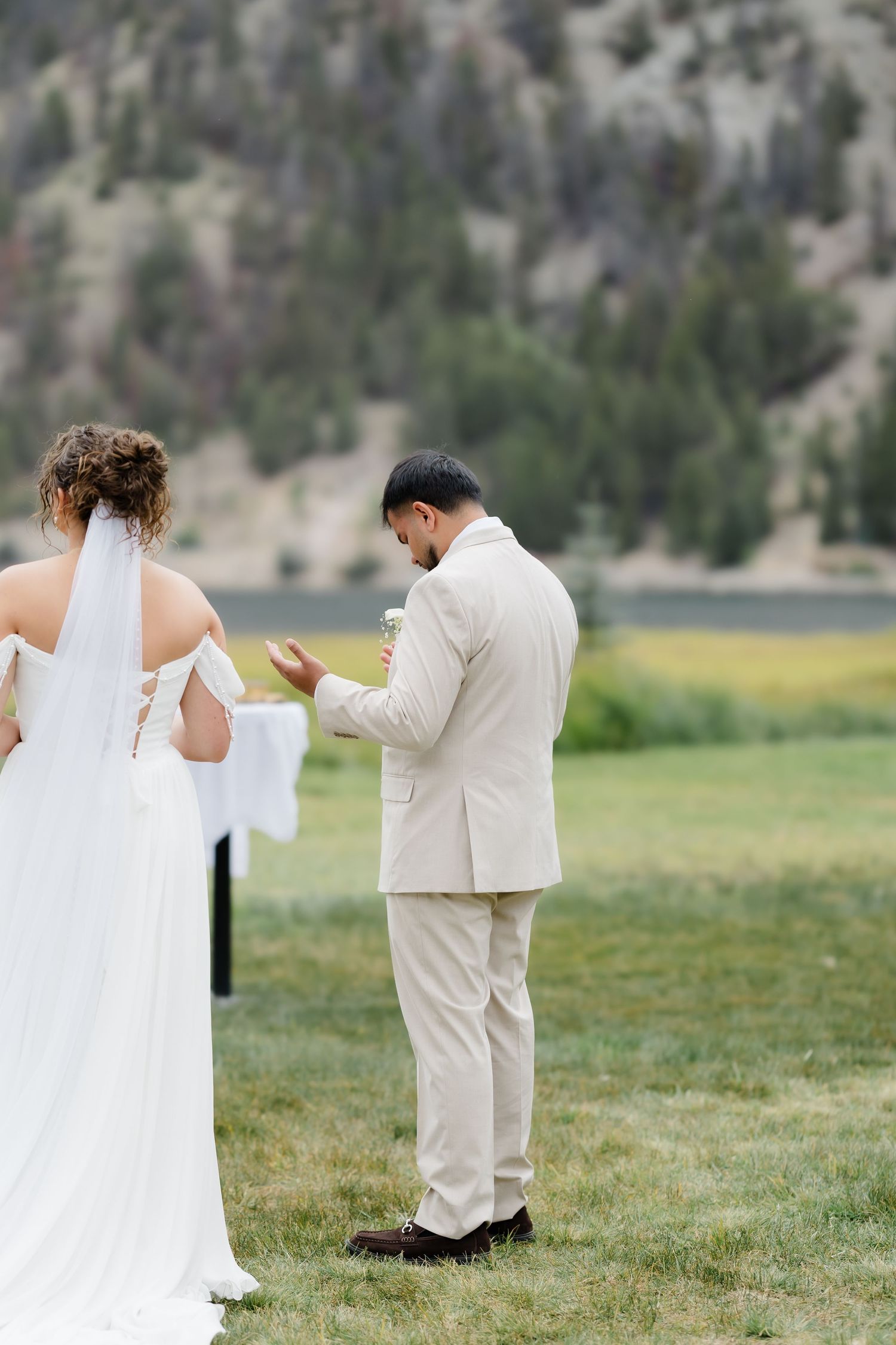 Groom in beige suit reads vows to bride in white dress during outdoor wedding ceremony with mountain backdrop.