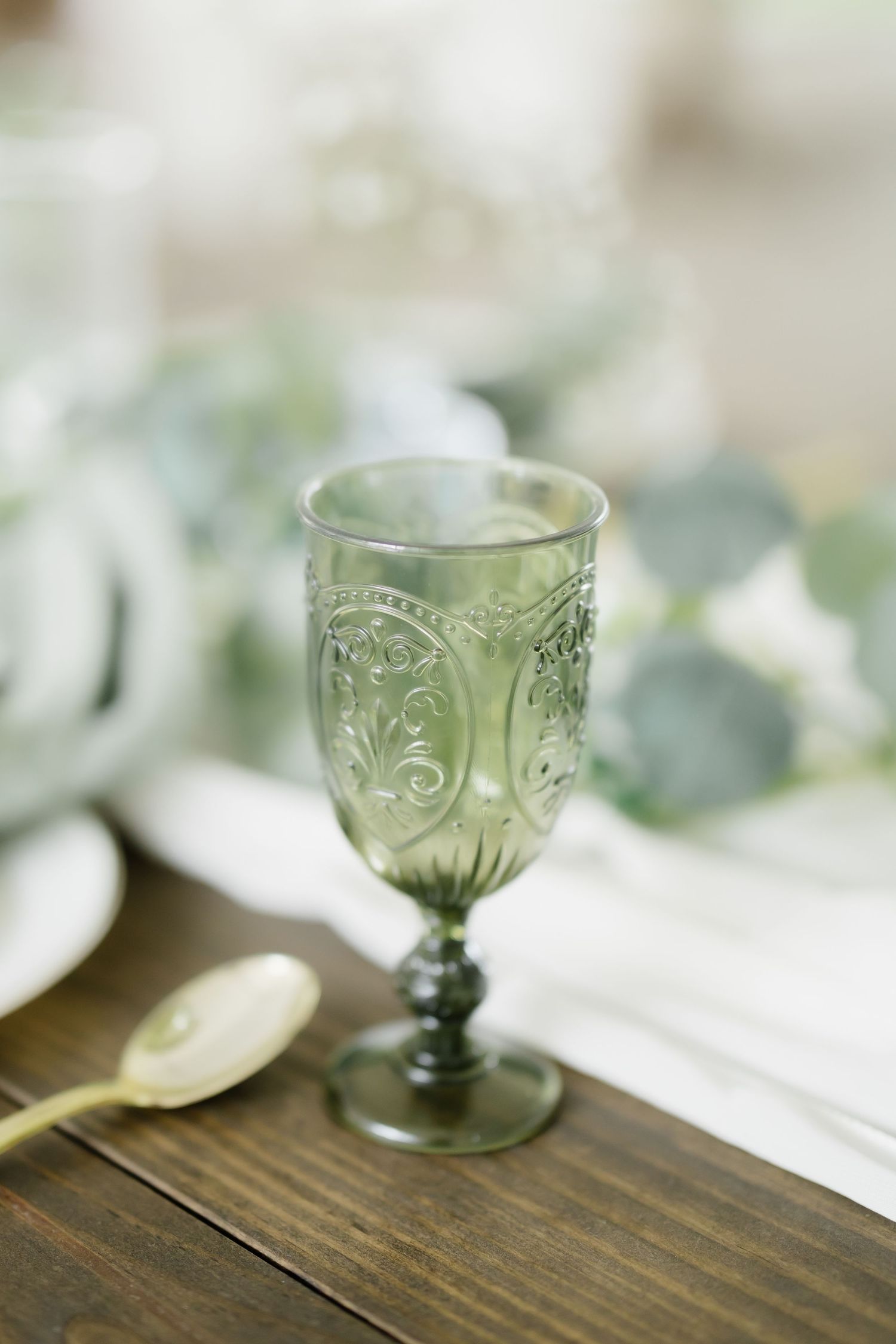 Elegant green embossed glass goblet on wooden table with eucalyptus and gold spoon at a wedding reception.