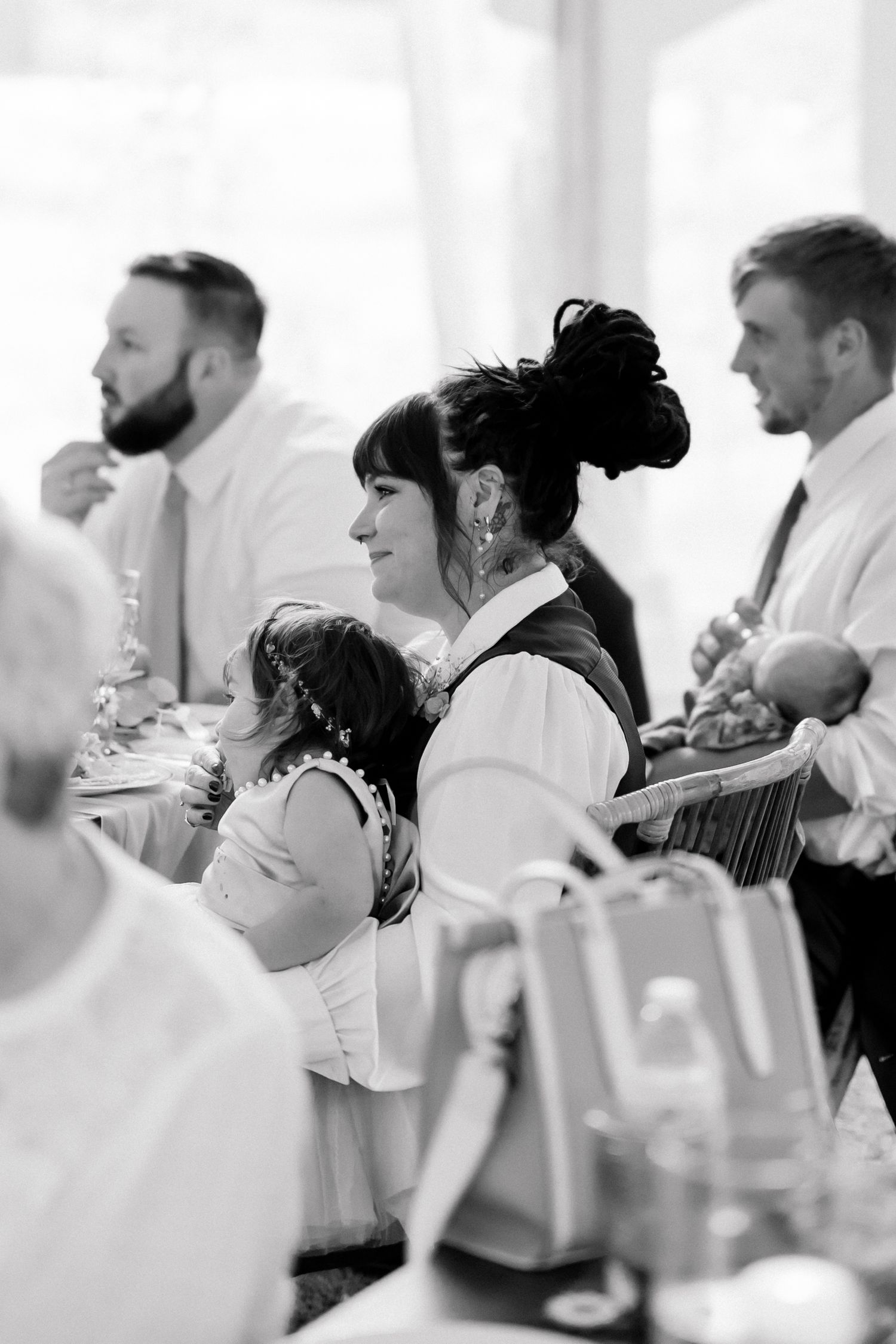 Black and white photo of wedding guests enjoying reception with woman holding child in foreground.