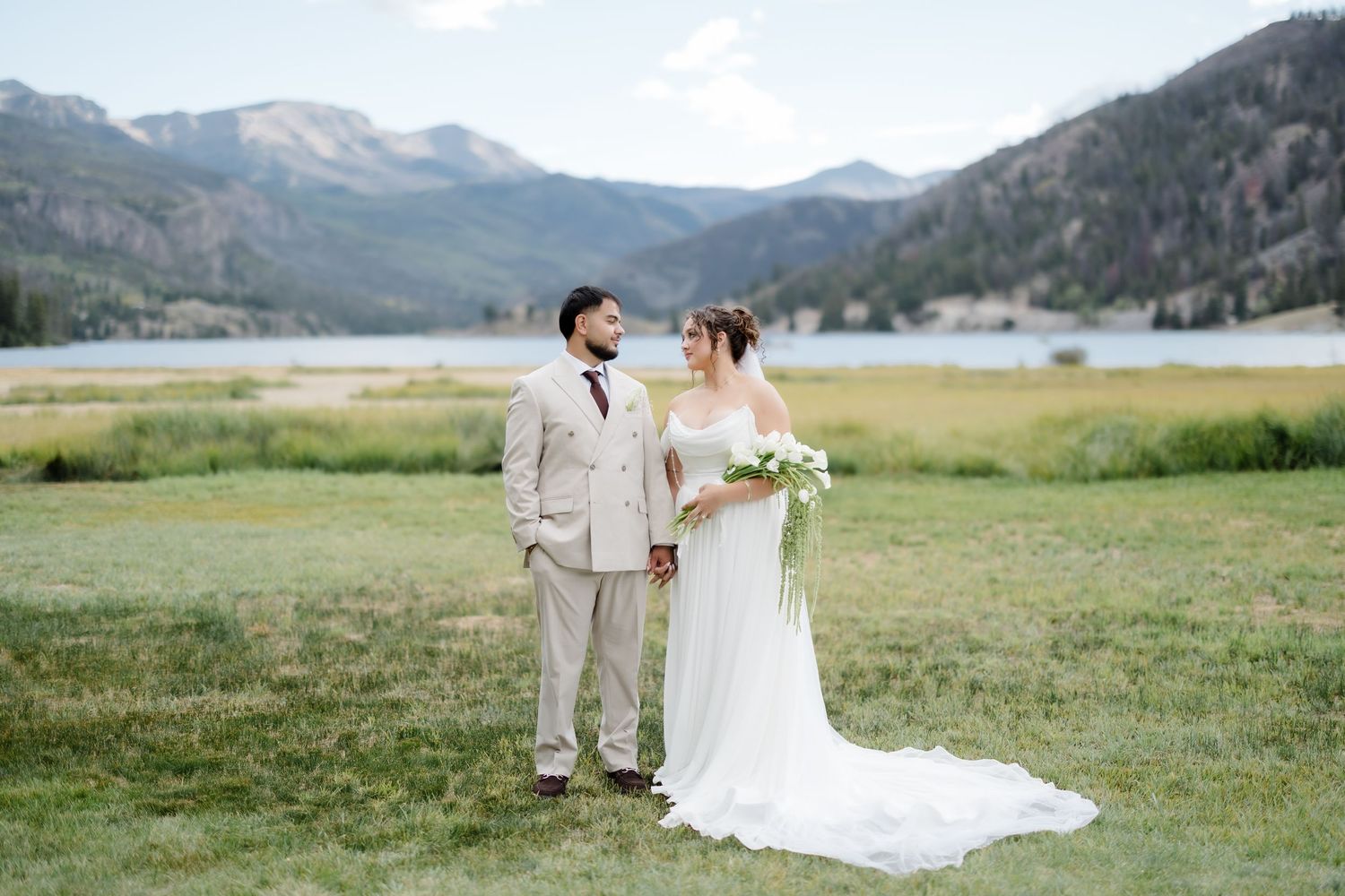 Bride and groom stand together in a mountain meadow with a lake and peaks backdrop on their wedding day.