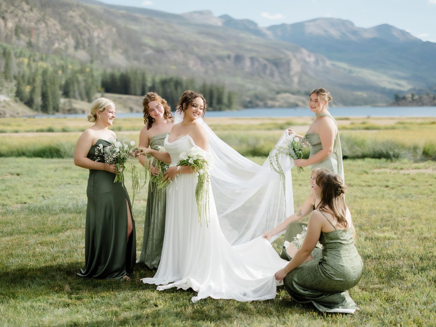Bride in white gown with bridesmaids in sage green dresses posing in mountain meadow near lake on wedding day.