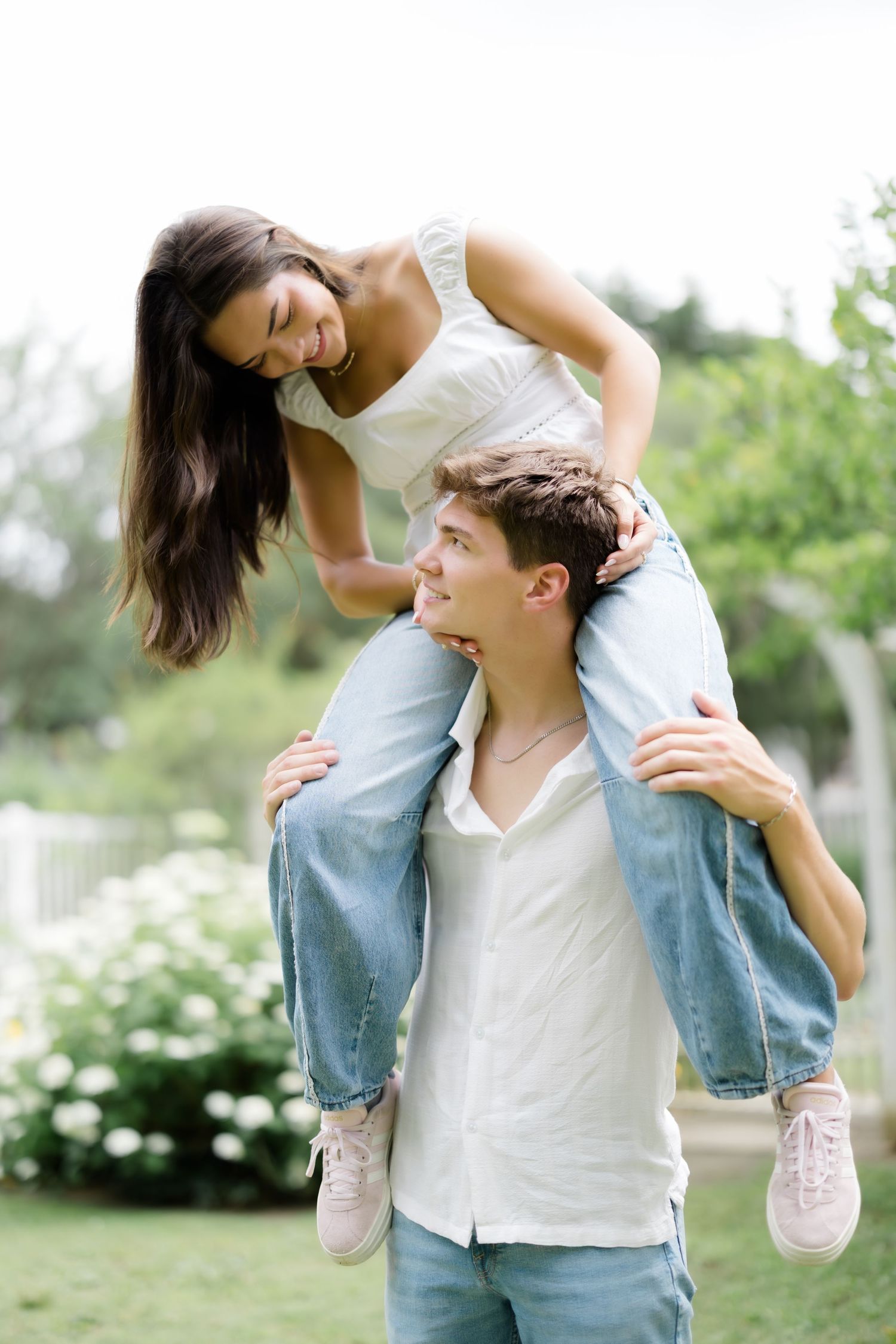 Young couple laughing outdoors, man giving woman a piggyback ride in a lush green garden setting.