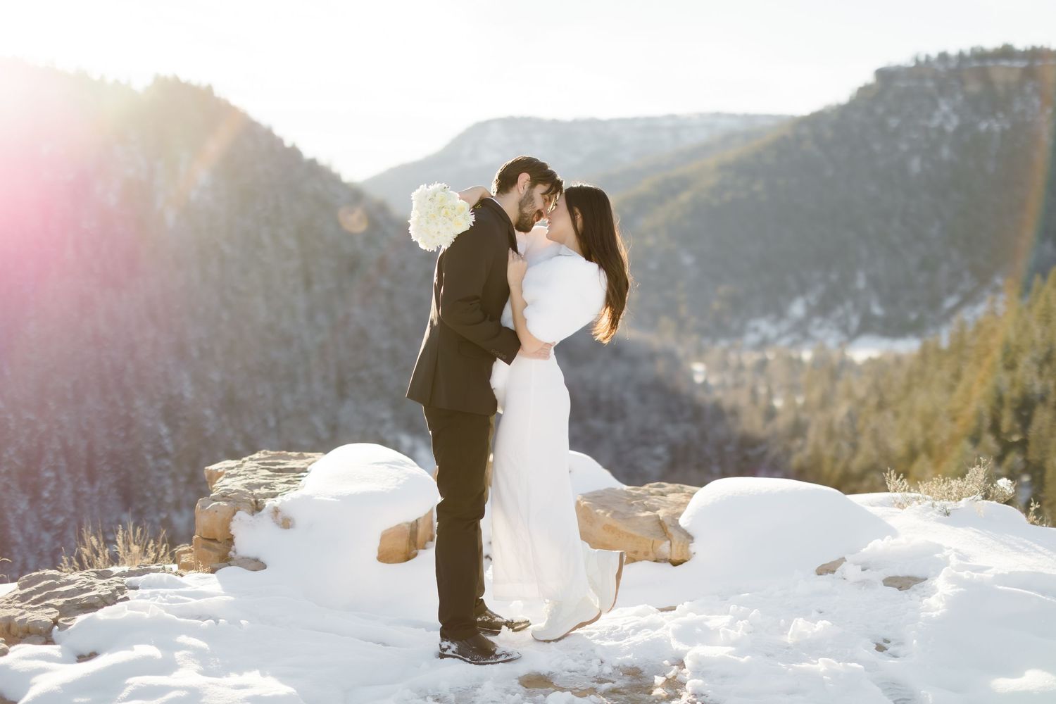 Bride and groom share a romantic kiss in snowy mountain landscape during winter wedding photo session.