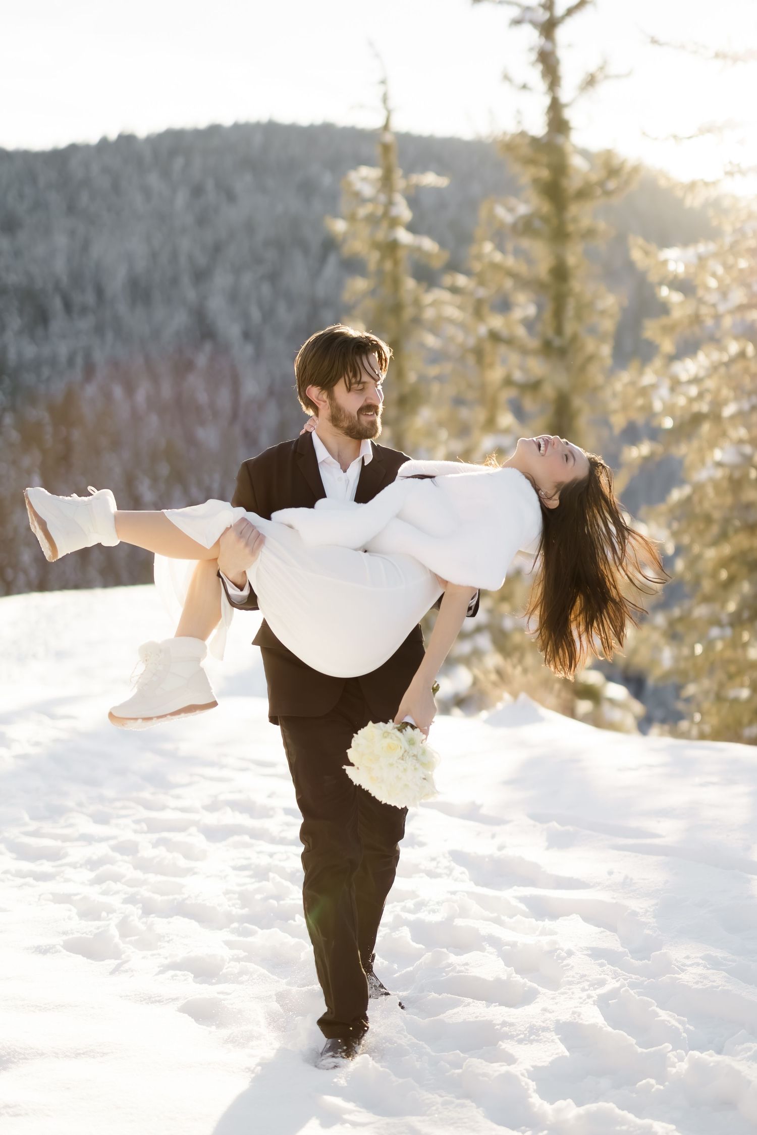 Laughing groom carries joyful bride in white dress through a sunny snowy mountain landscape.