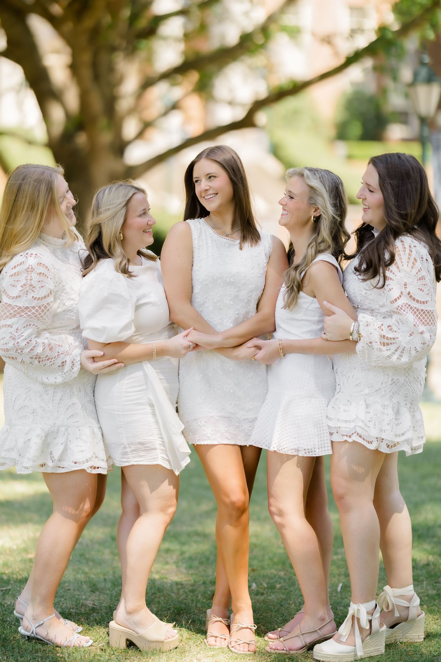 Five women in white lace dresses pose together outdoors, smiling and laughing in a sunny park setting.