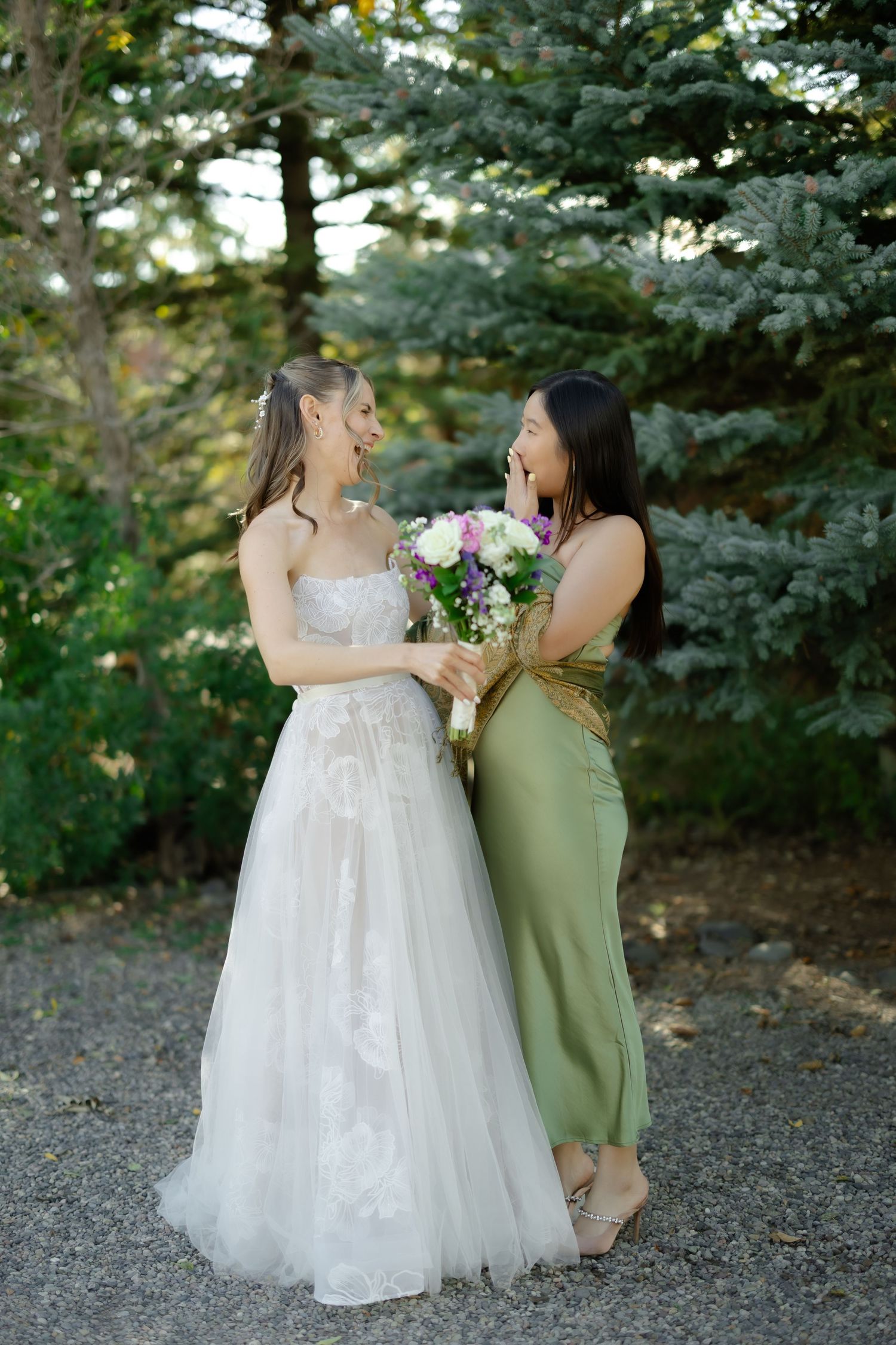 Bride in white gown and bridesmaid in green dress share joyful moment holding bouquet outdoors near pine trees.