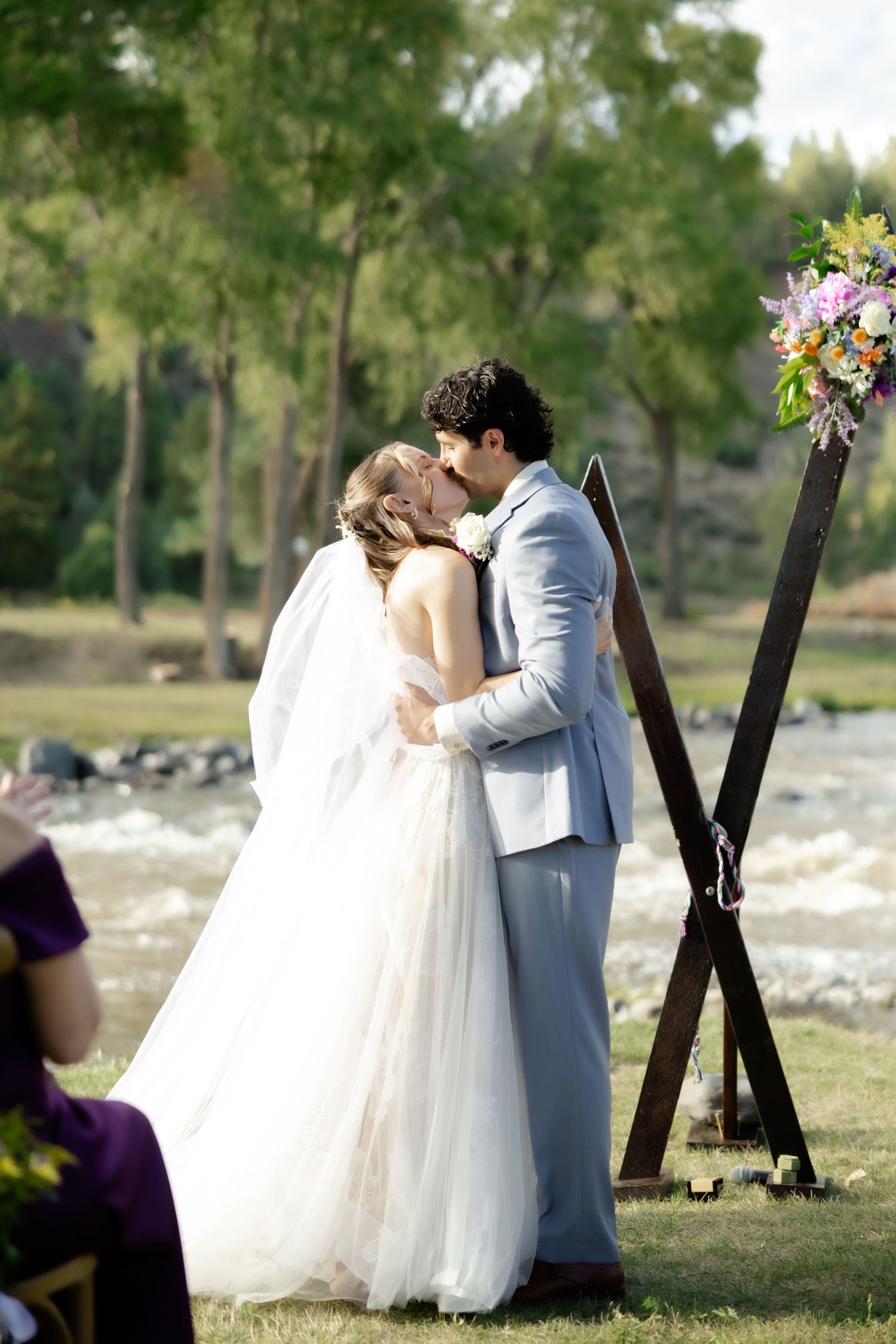 Bride and groom share their first kiss at an outdoor wedding ceremony beside a rustic wooden arch.