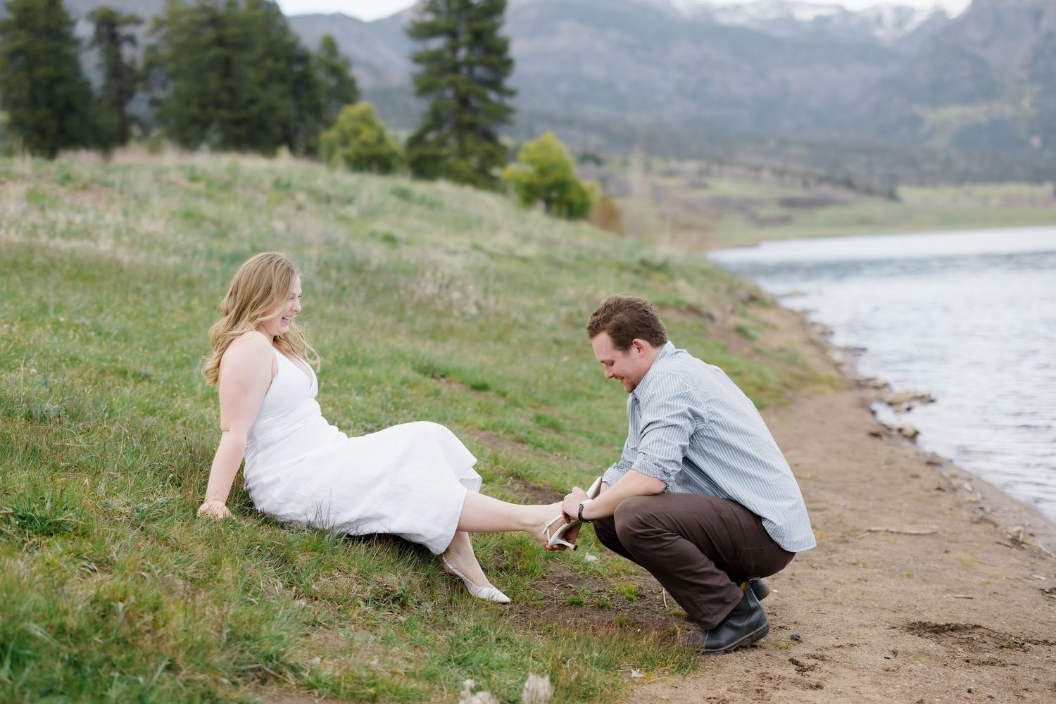 Man kneeling by lakeside helping woman with her shoes as she sits on grassy bank near water.