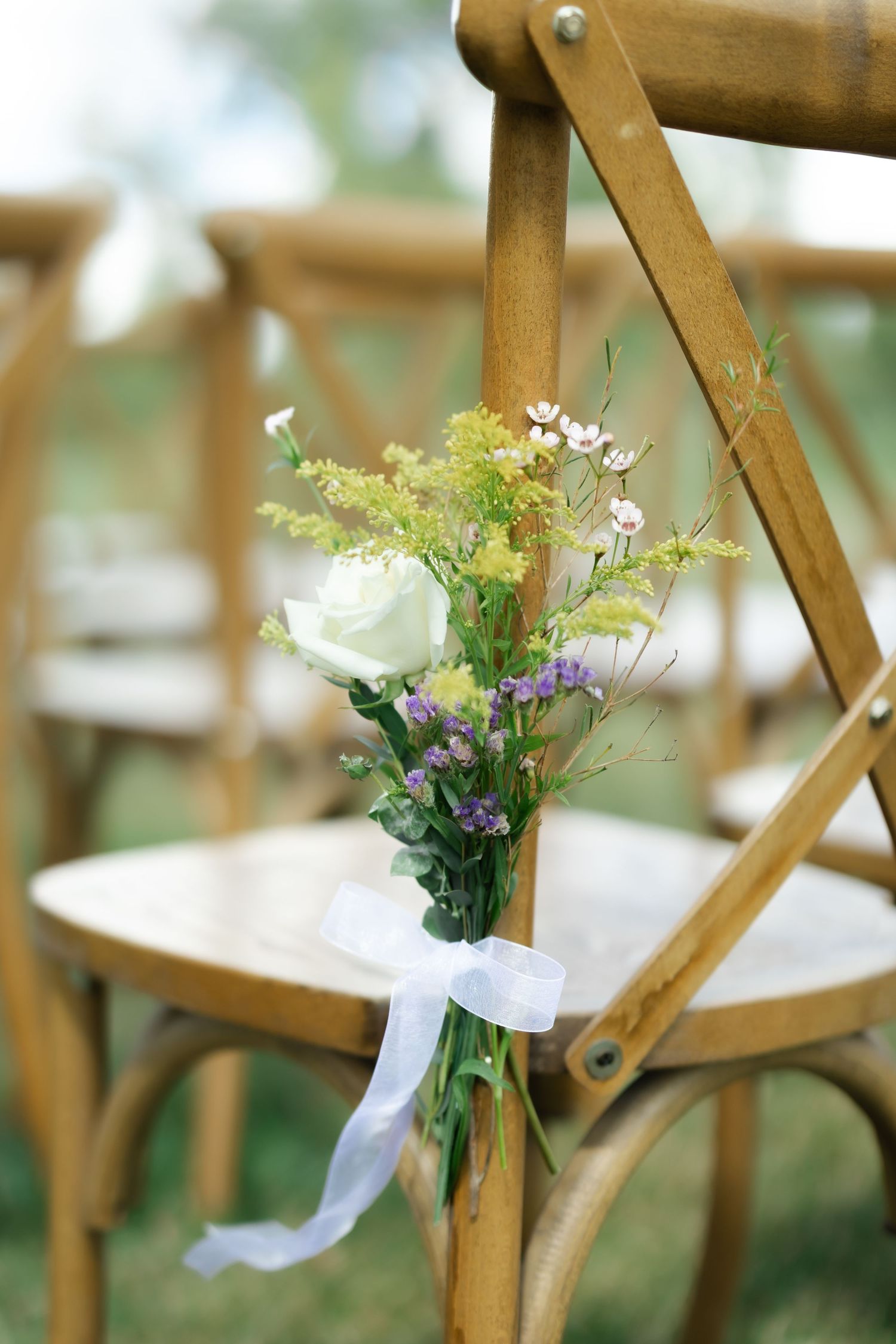 White rose and wildflower bouquet tied with ribbon on wooden cross-back chair at outdoor wedding ceremony.