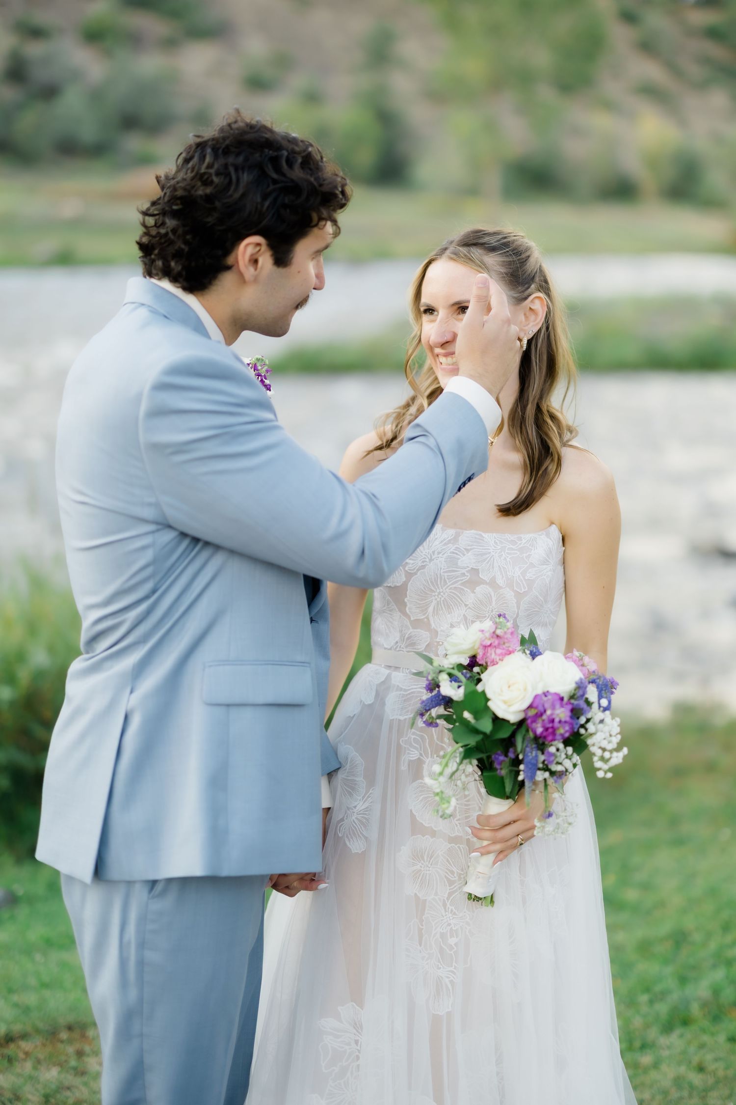 Groom in light blue suit gently touches bride's face as she holds purple and white bouquet outdoors.