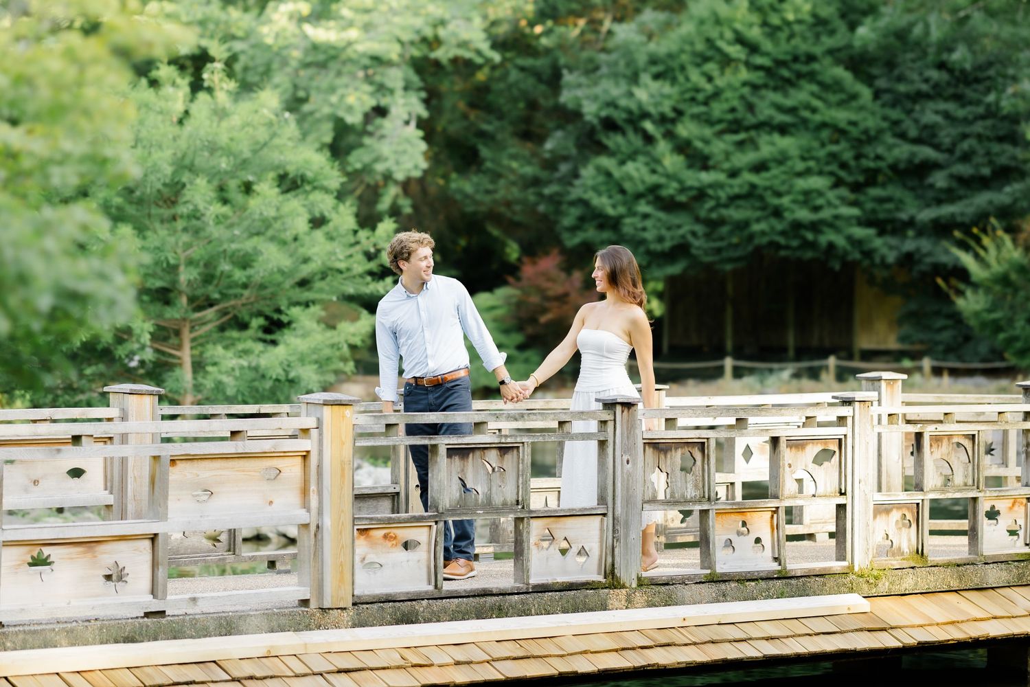 Couple holding hands on a wooden bridge surrounded by lush green trees in an outdoor engagement photo.