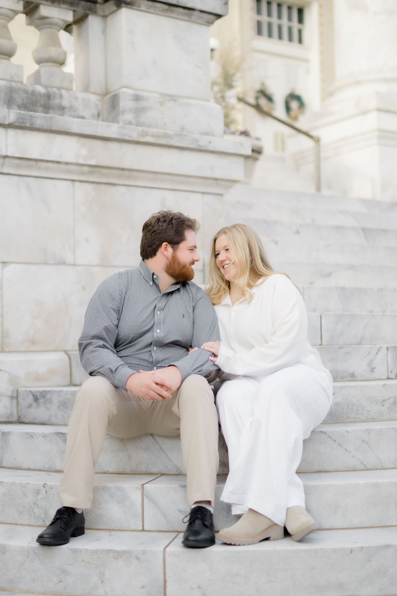 Couple sitting on marble steps, smiling at each other during an engagement photo session.