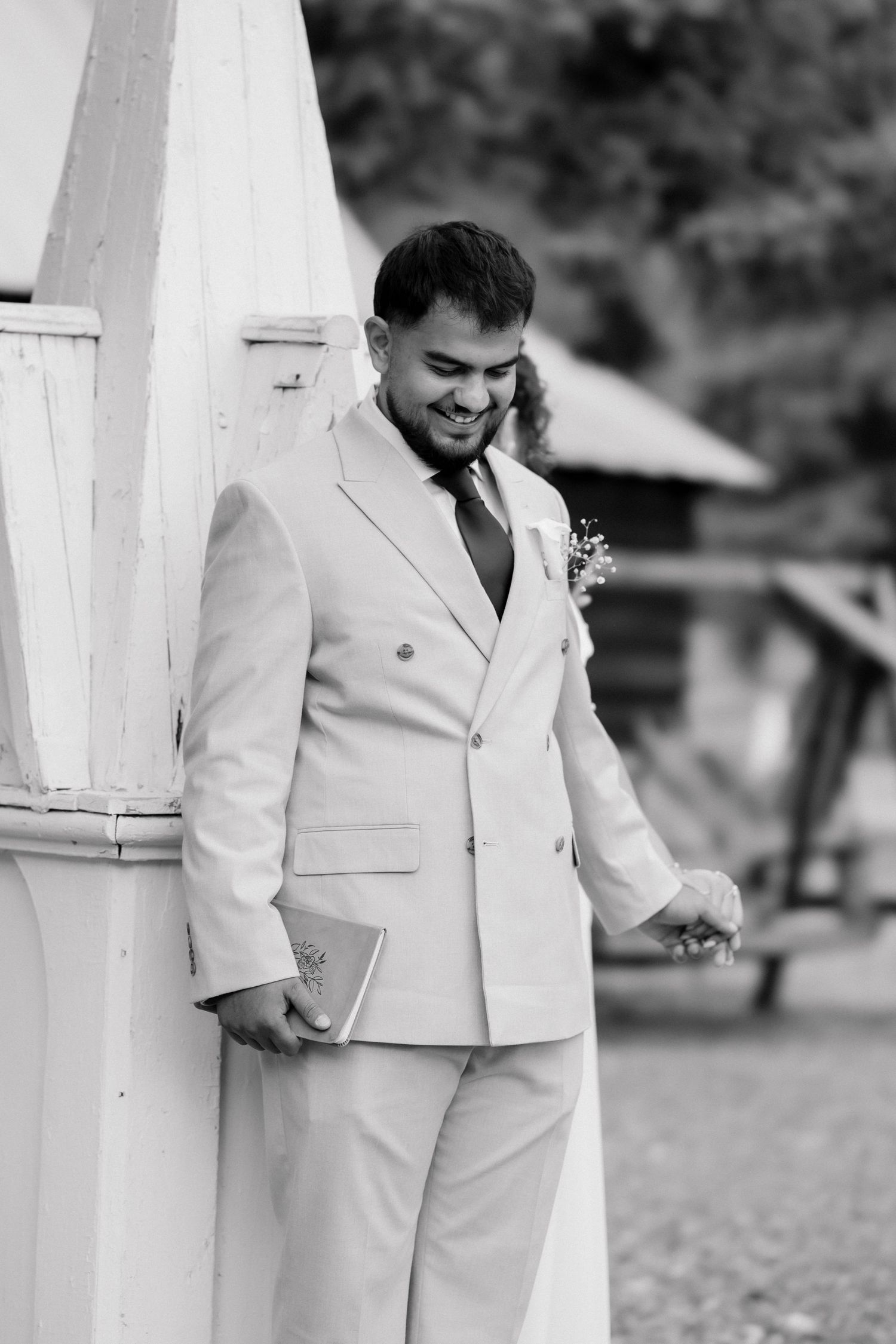 Black and white photo of a smiling groom in a white suit with boutonniere, holding a book outdoors.