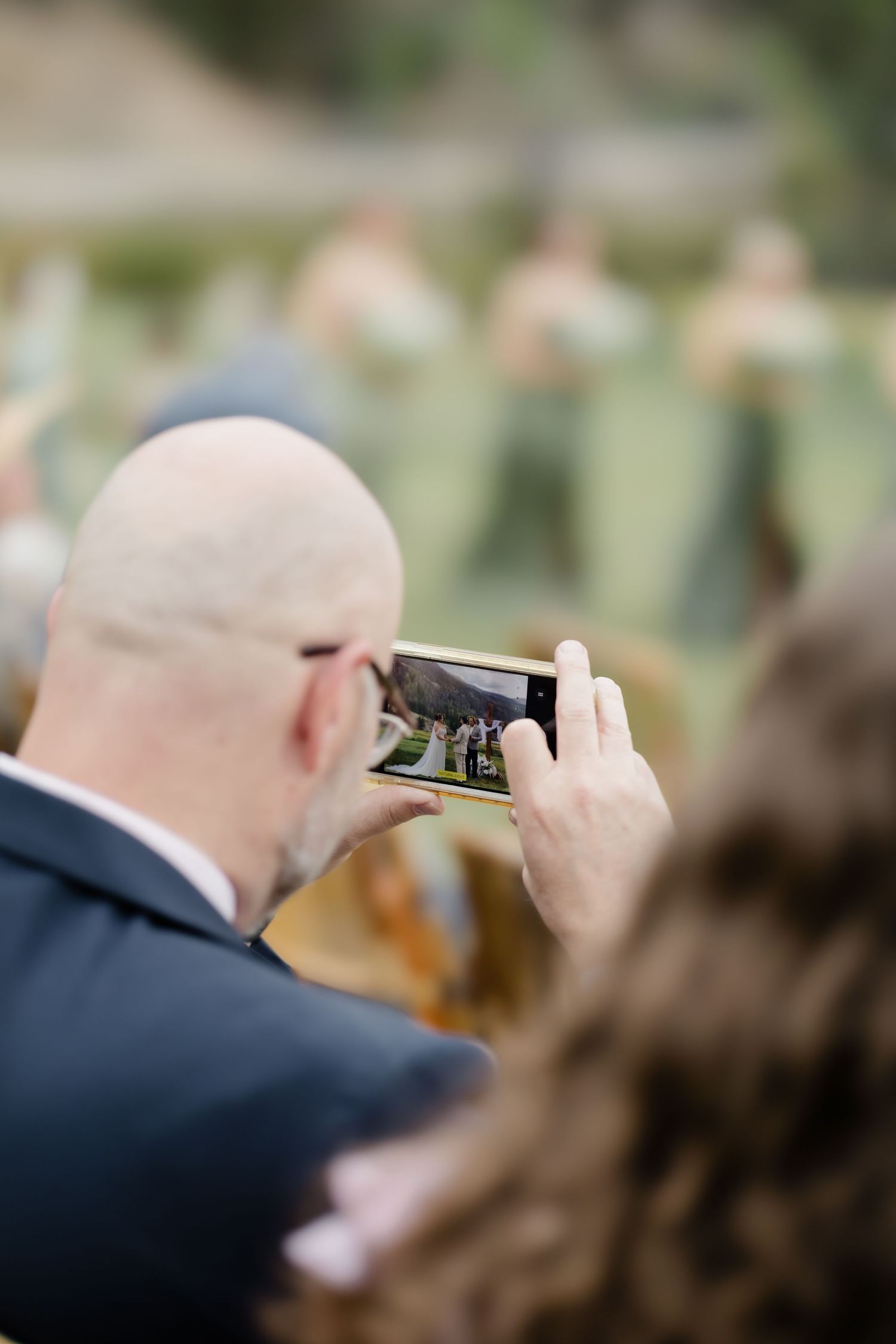 Bald wedding guest photographs outdoor ceremony on smartphone at elegant garden wedding.