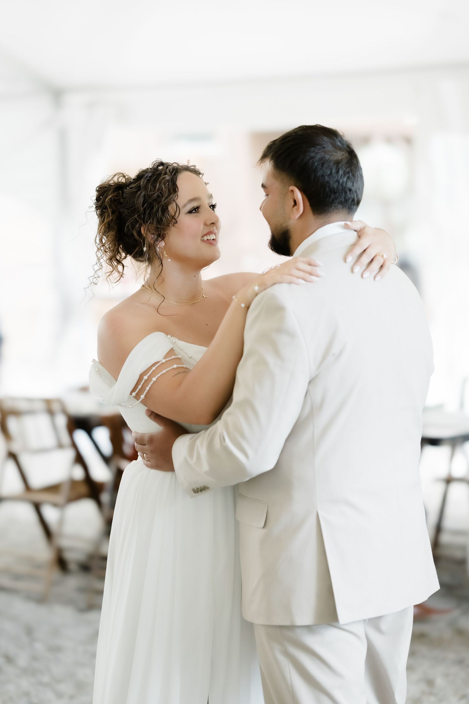 Bride and groom sharing first dance, smiling at each other in elegant white wedding attire.