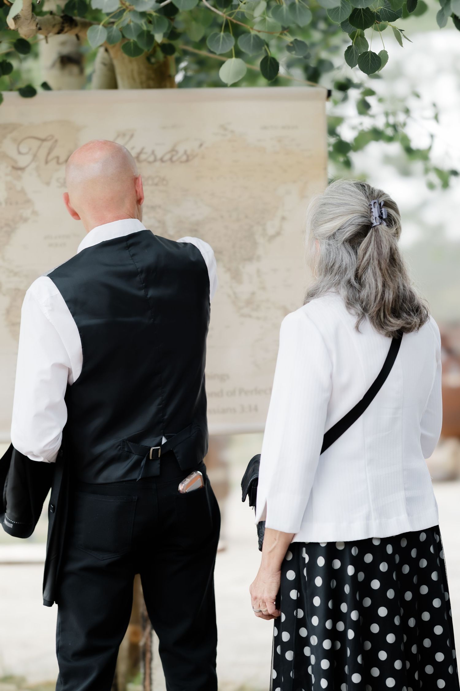 Couple viewed from behind examining a vintage world map display outdoors, man in vest and woman in polka dot skirt.