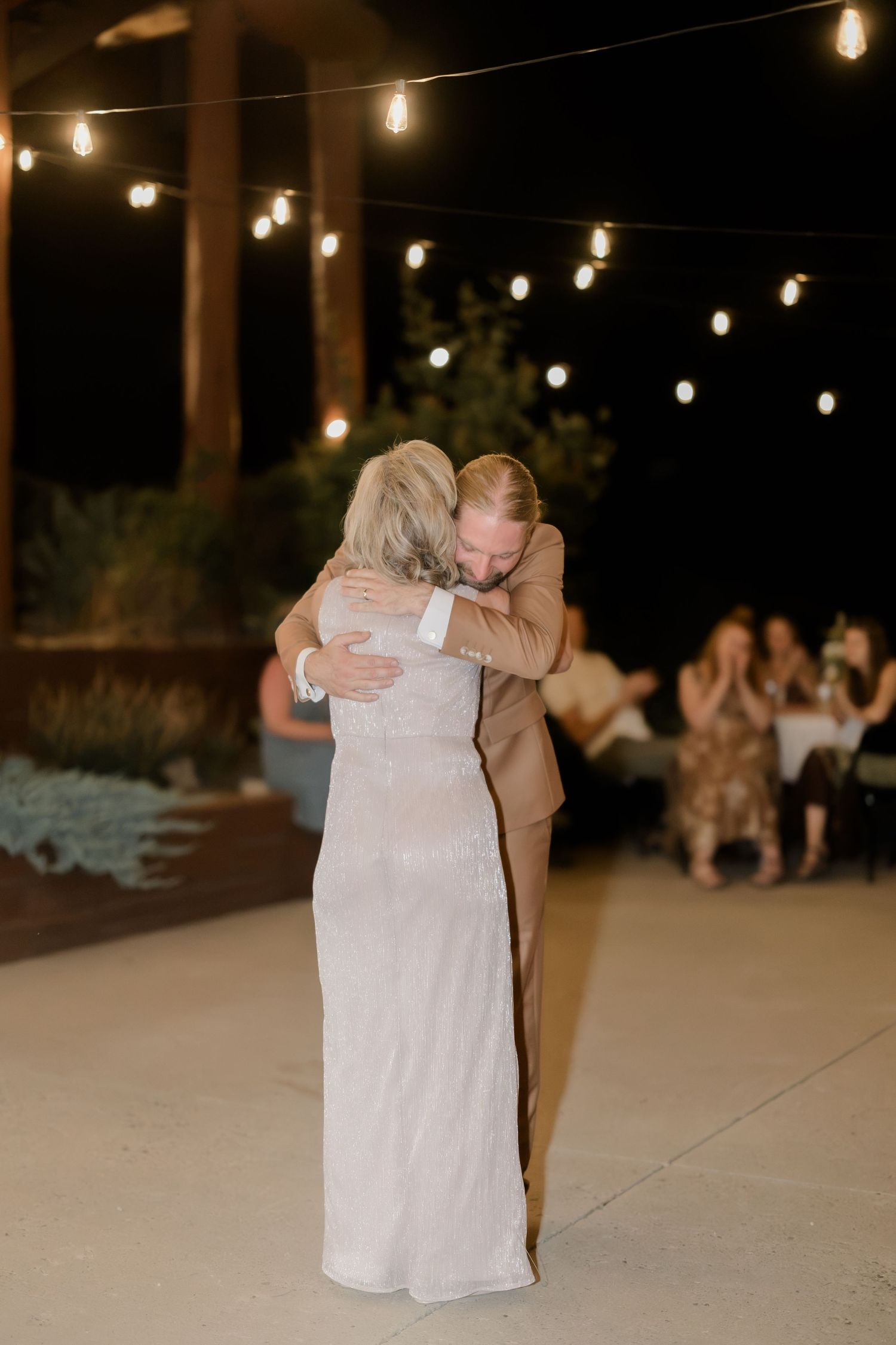 A groom in a tan suit shares an emotional dance with his mother under string lights at an outdoor wedding reception.